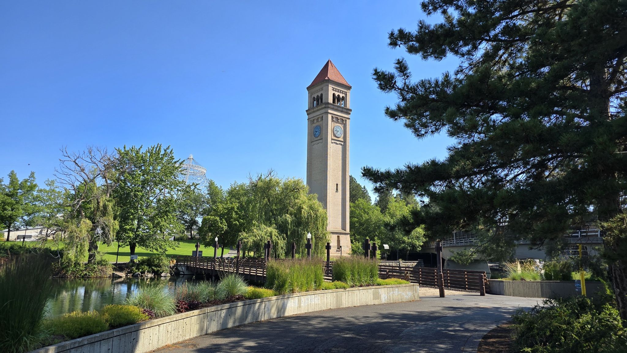 Tall clock tower in a green park with a pond and trees on a sunny day. - Garbage Goat Sculpture – Riverfront Park loop from Spokane