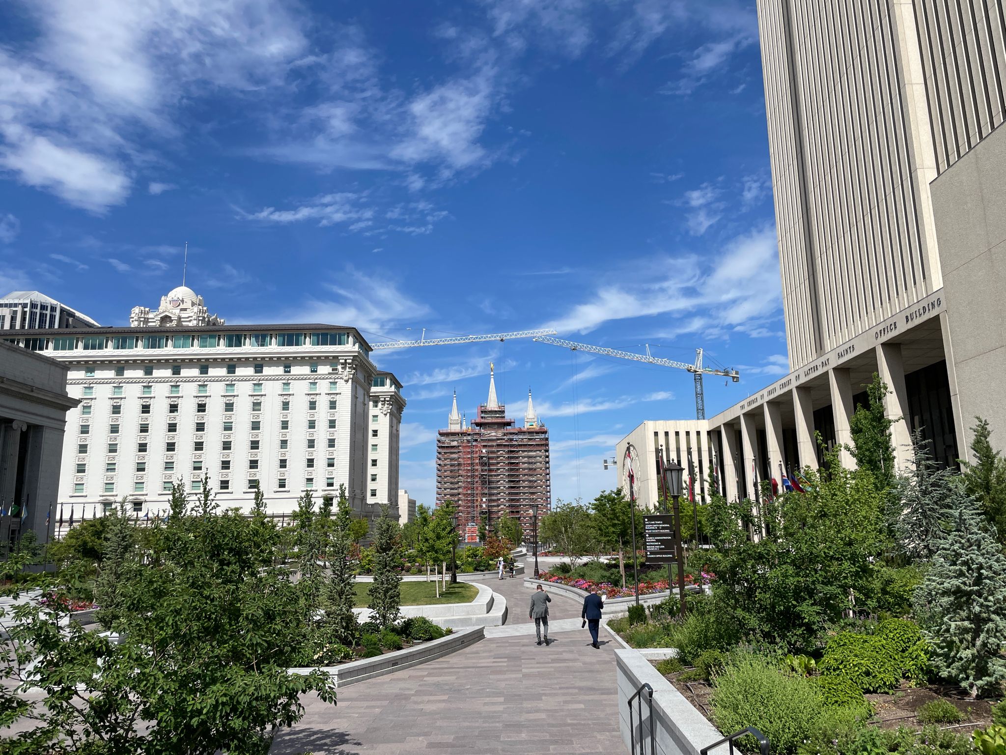 Daytime cityscape with tall buildings, green trees, and people walking on a paved path. - Cycle the City