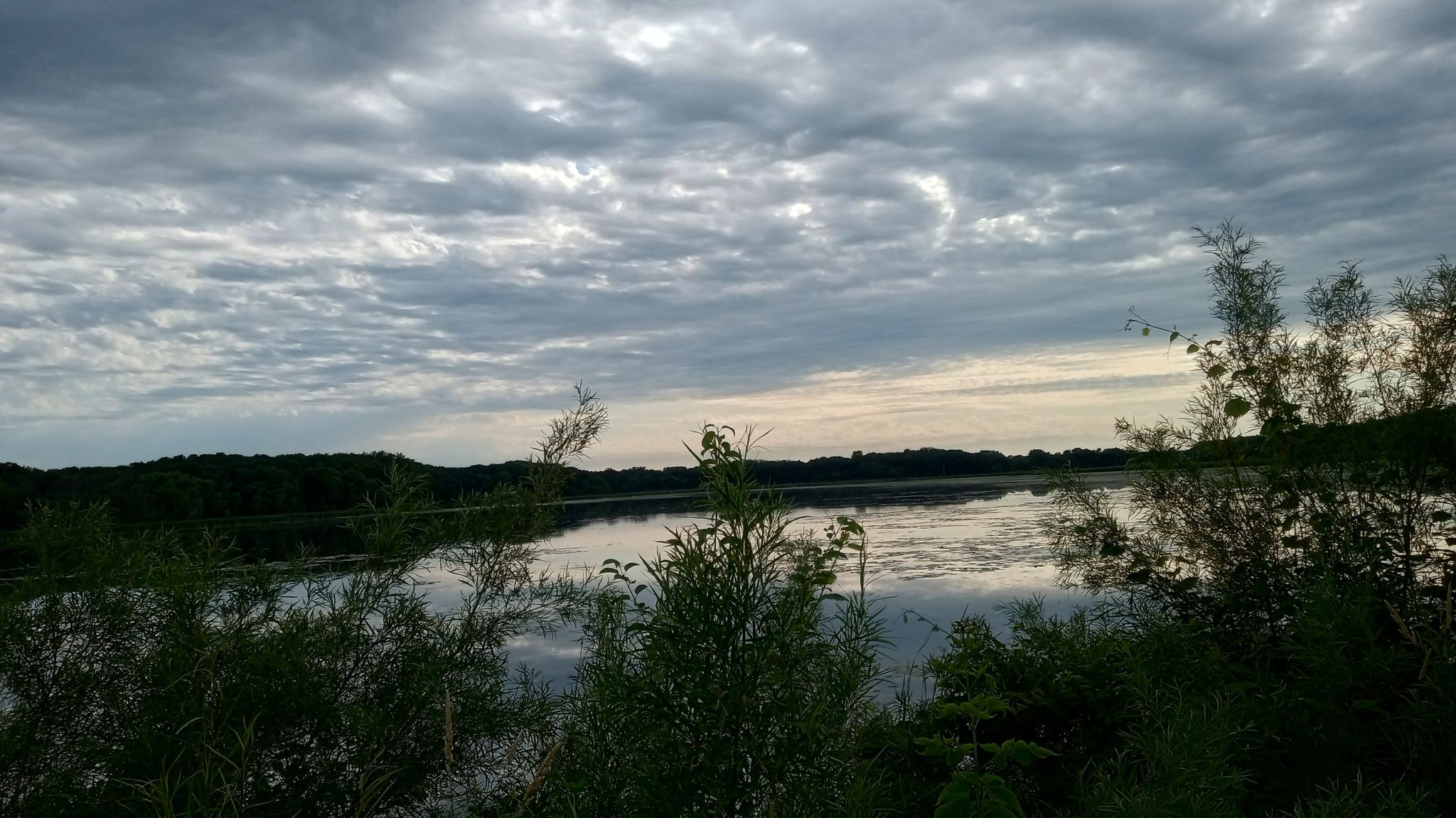 A calm lake reflecting a dramatic cloudy sky at dusk, with dark foliage. - Biking/walking bridge over 212 – Riley Lake Park loop from Chanhassen