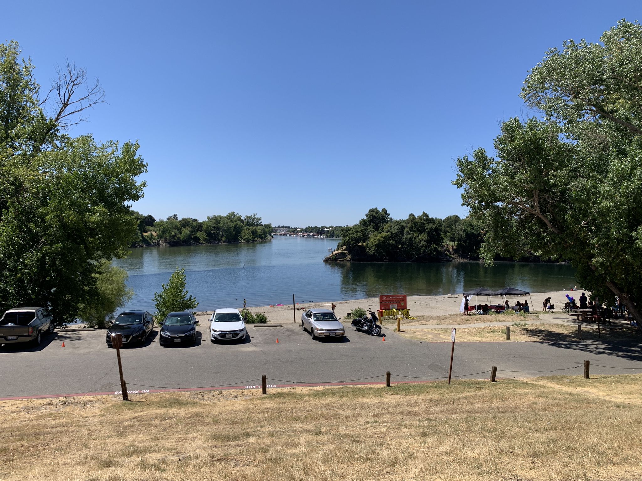Sunny riverfront park with sandy beach, parked cars, and people. - Two Rivers and Downtown Sacramento Loop