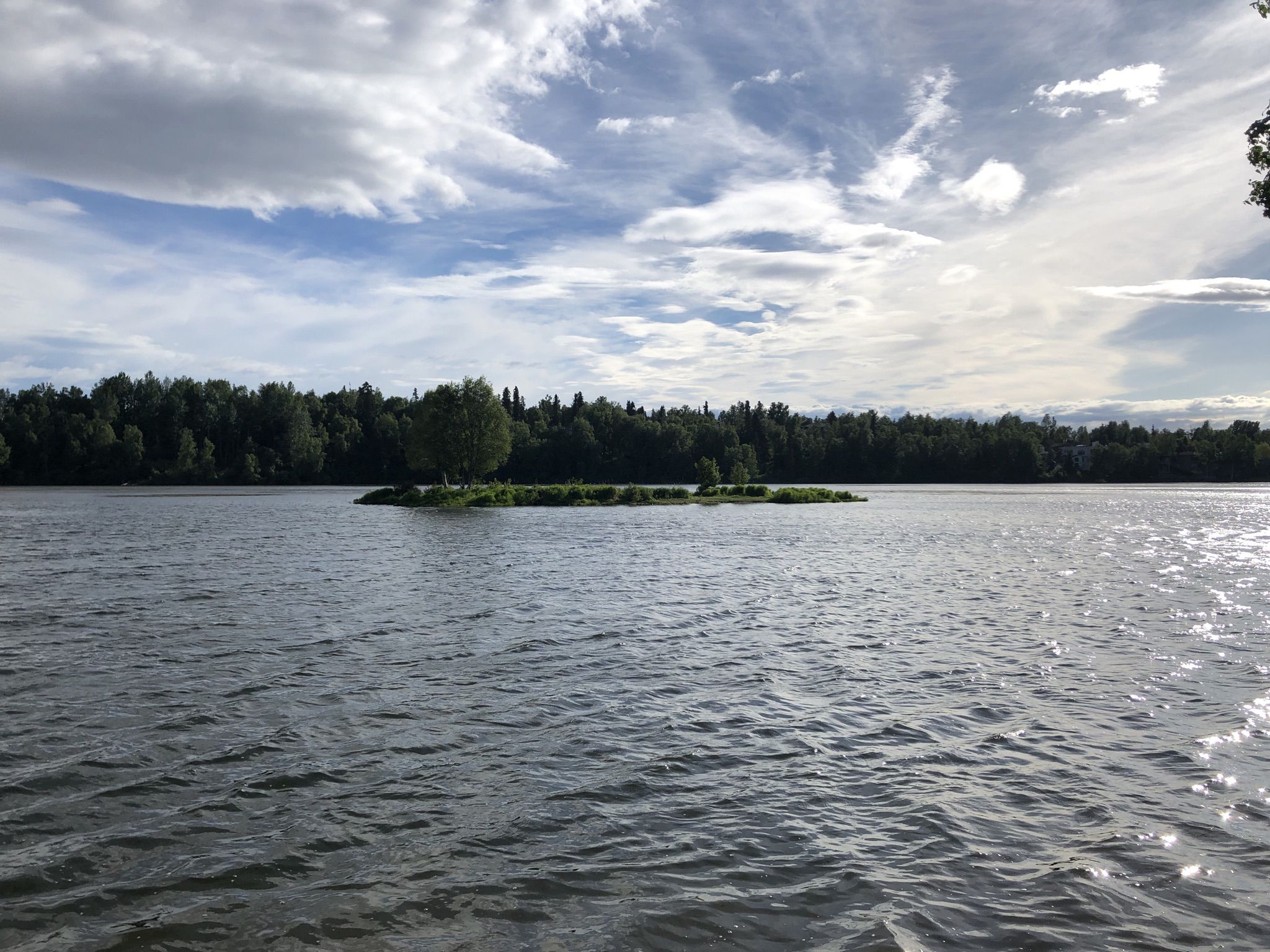 Wide lake with a small forested island under a bright, partly cloudy sky. - Chester Creek Trail