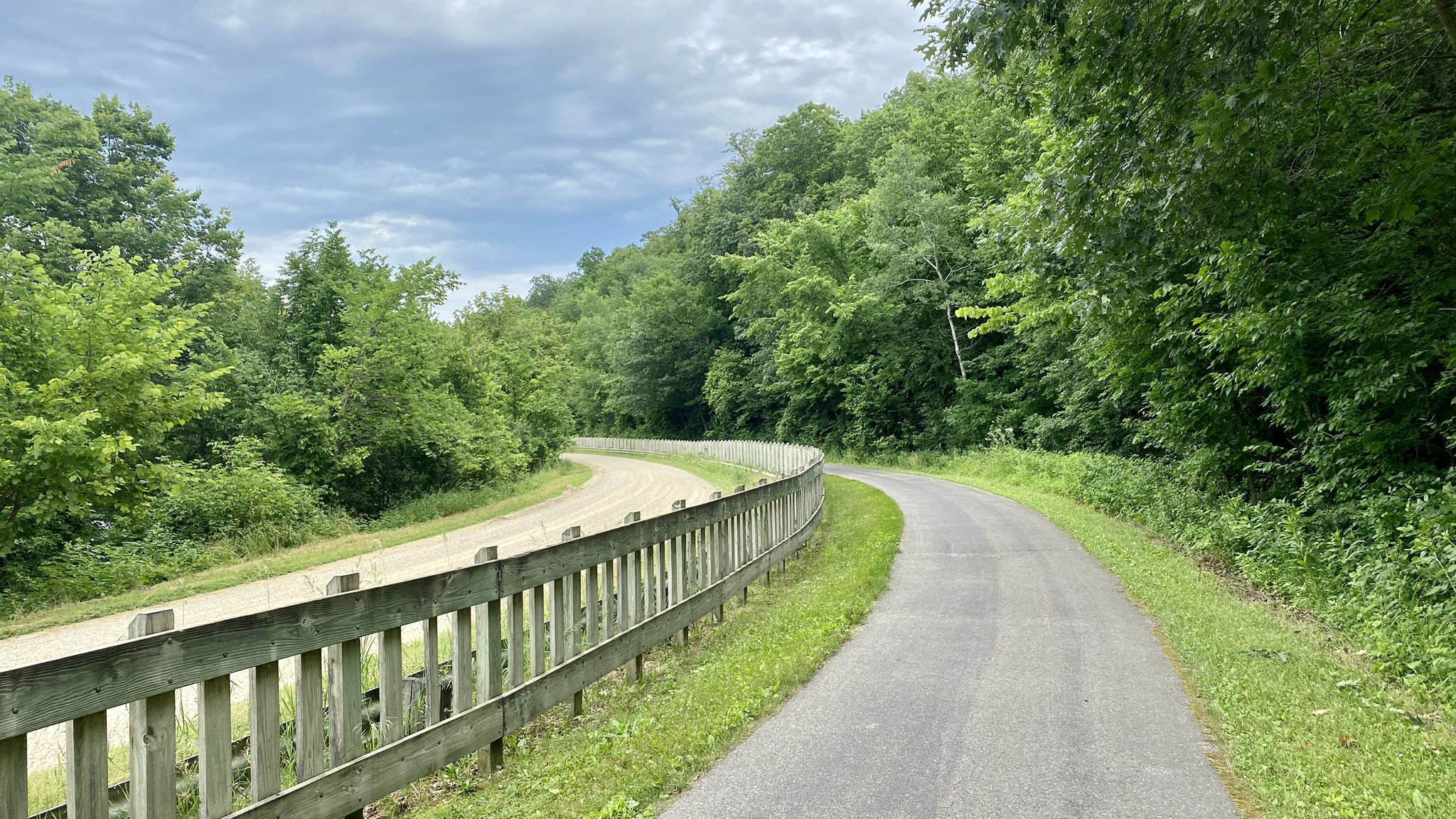A paved path and dirt path with a fence, surrounded by green trees. - Welch Village – M80 - Train Wreck Site loop from Welch
