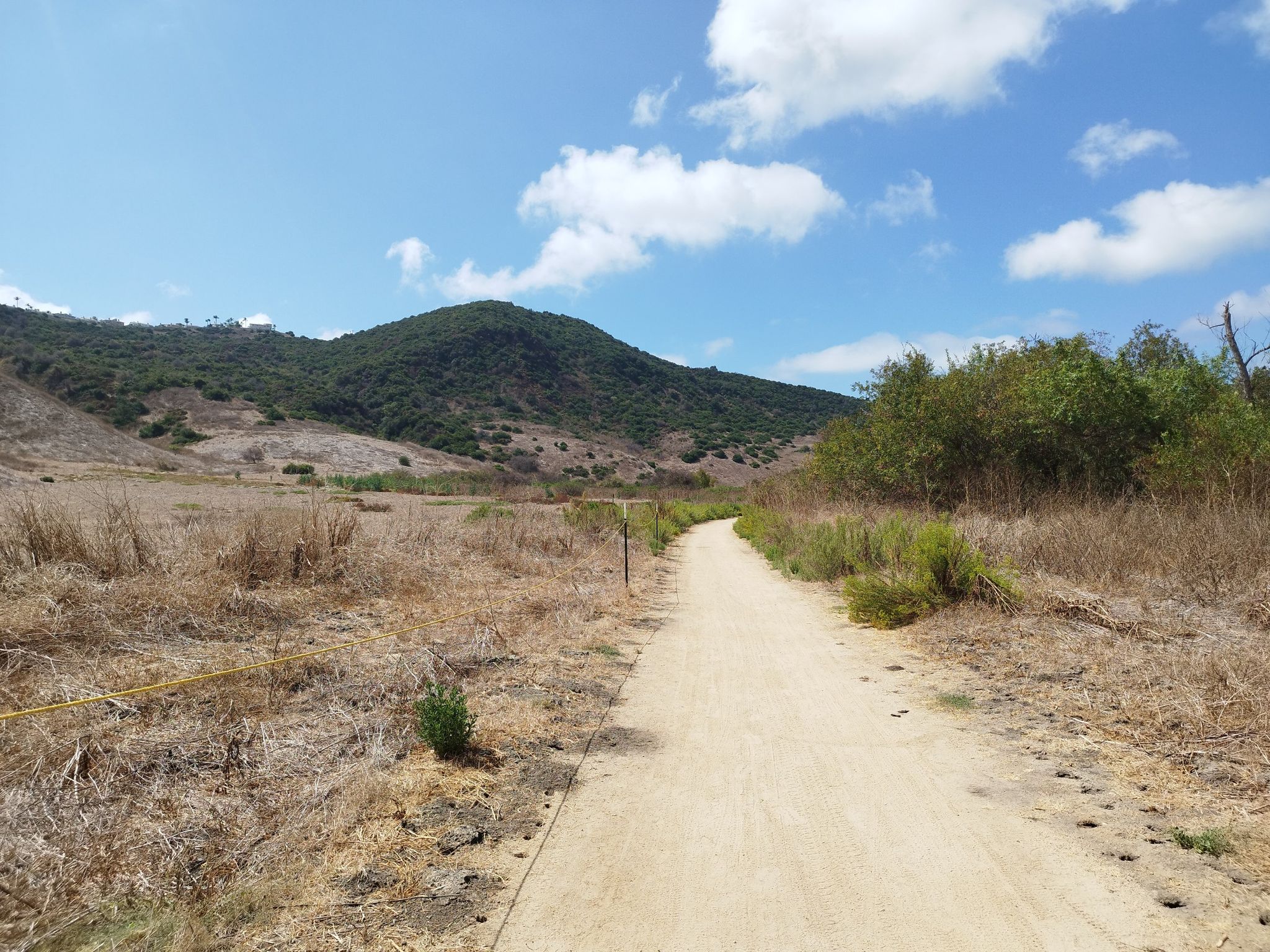 A dirt path winds through dry fields towards green hills under a sunny sky. - Aliso Creek Bikeway Loop