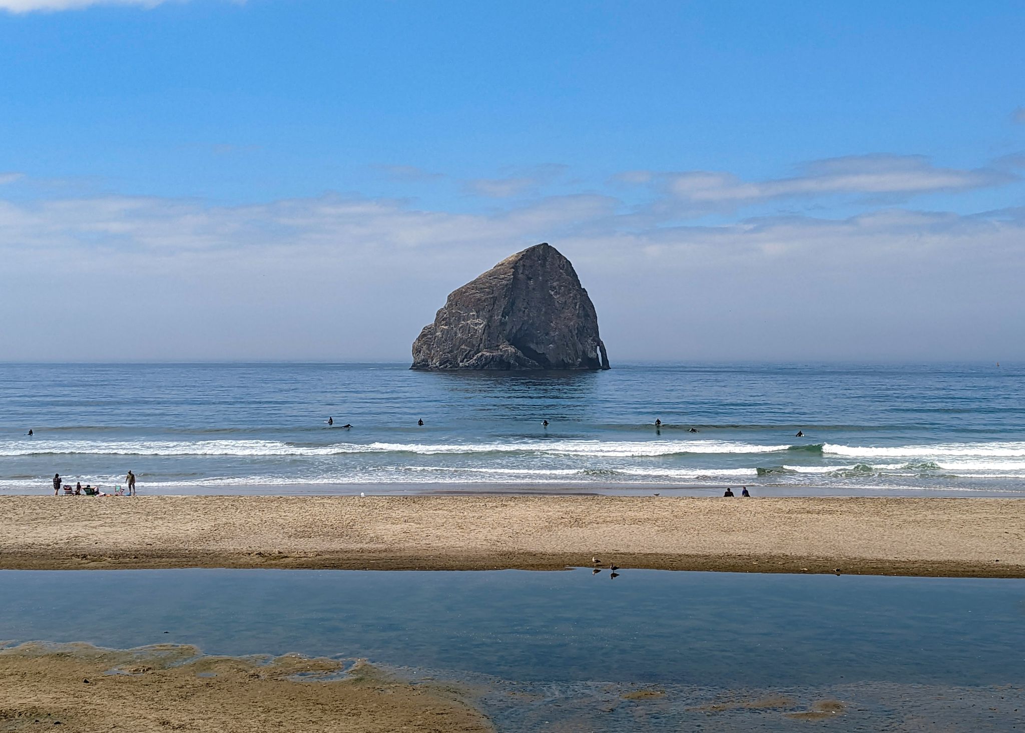 Large sea stack in ocean, surfers and people on sandy beach, bright day. - Pacific City loop