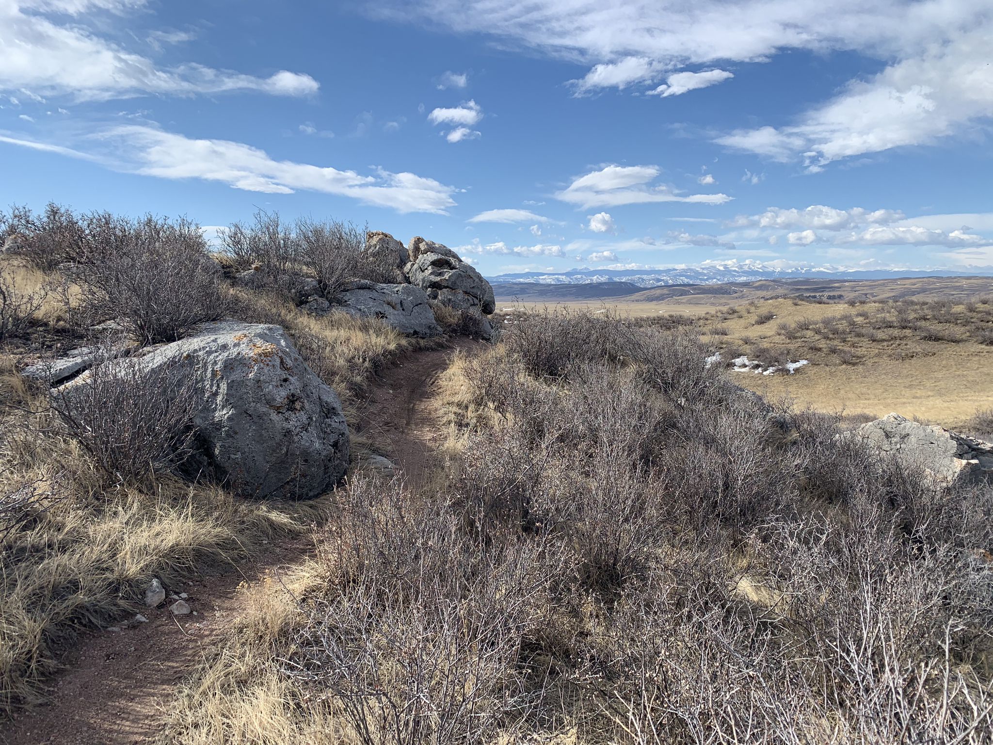A dirt path winds past large rocks and dry bushes under a blue sky. - Horse water tank loop from Lindenmeier Overlook