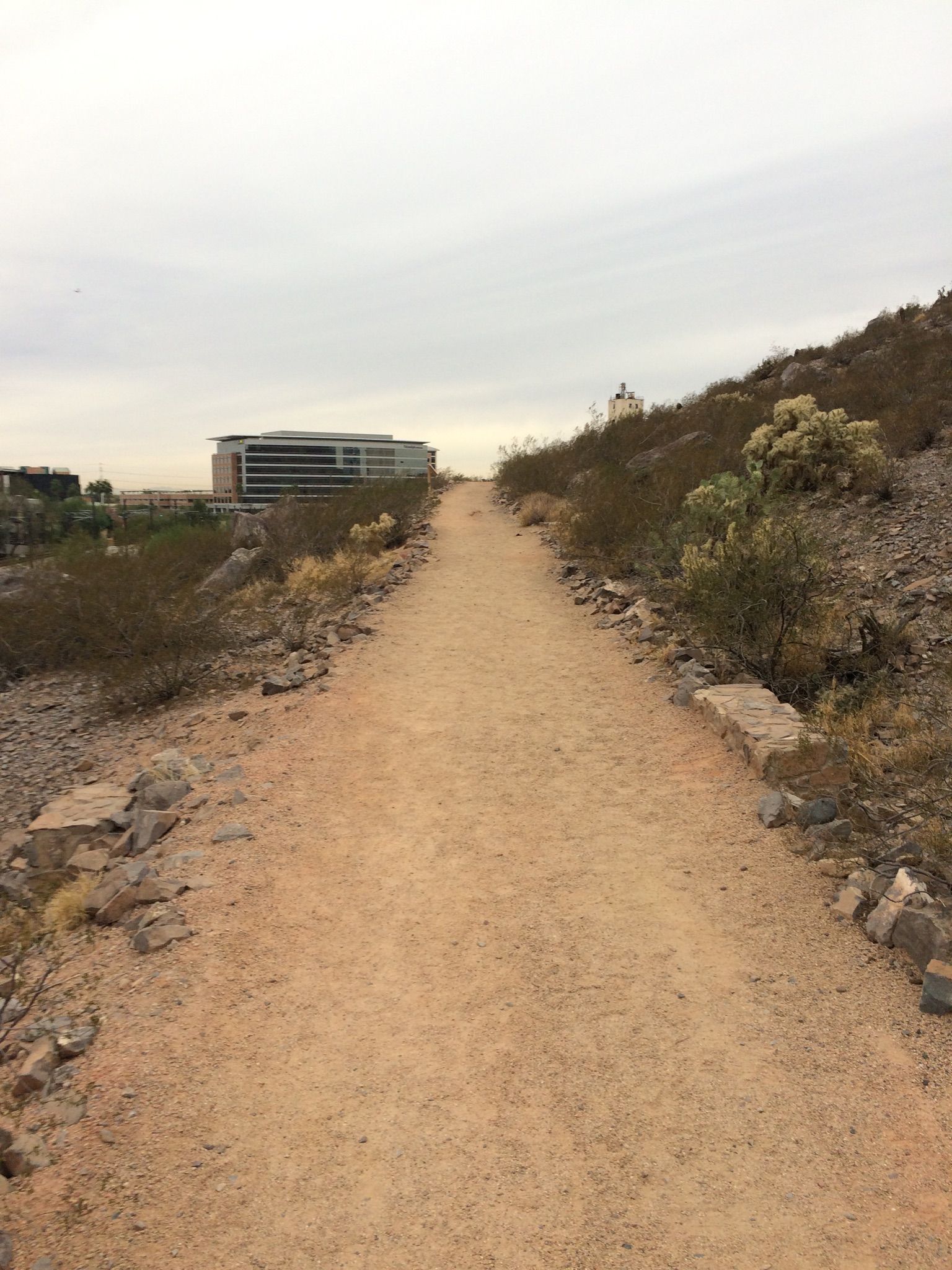 A dirt path winds through arid hills towards a distant building under an overcast sky. - Leonard Monti Trail