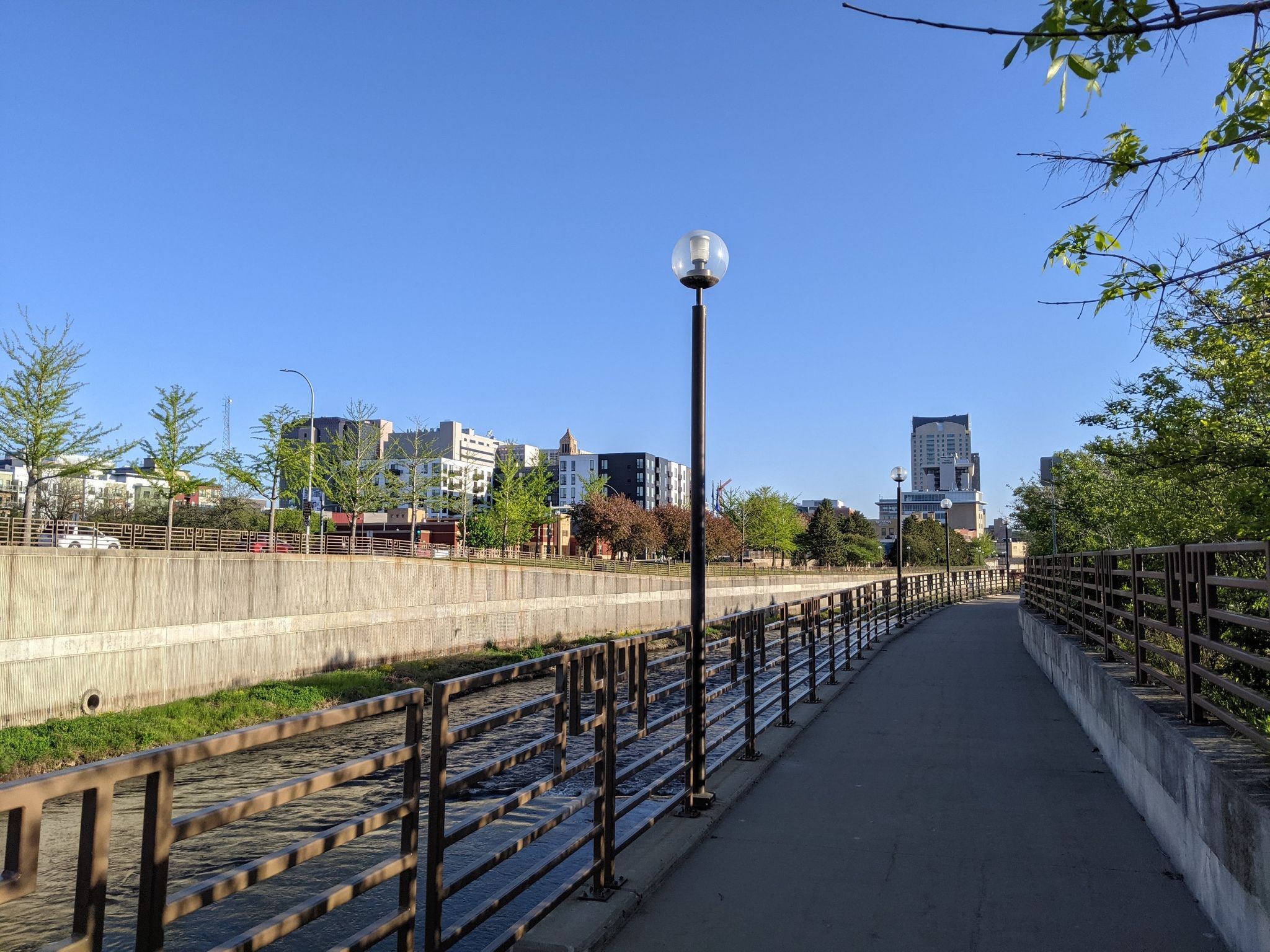 Paved path next to a river, city buildings in distance, under clear blue sky. - Zumbro River Trail