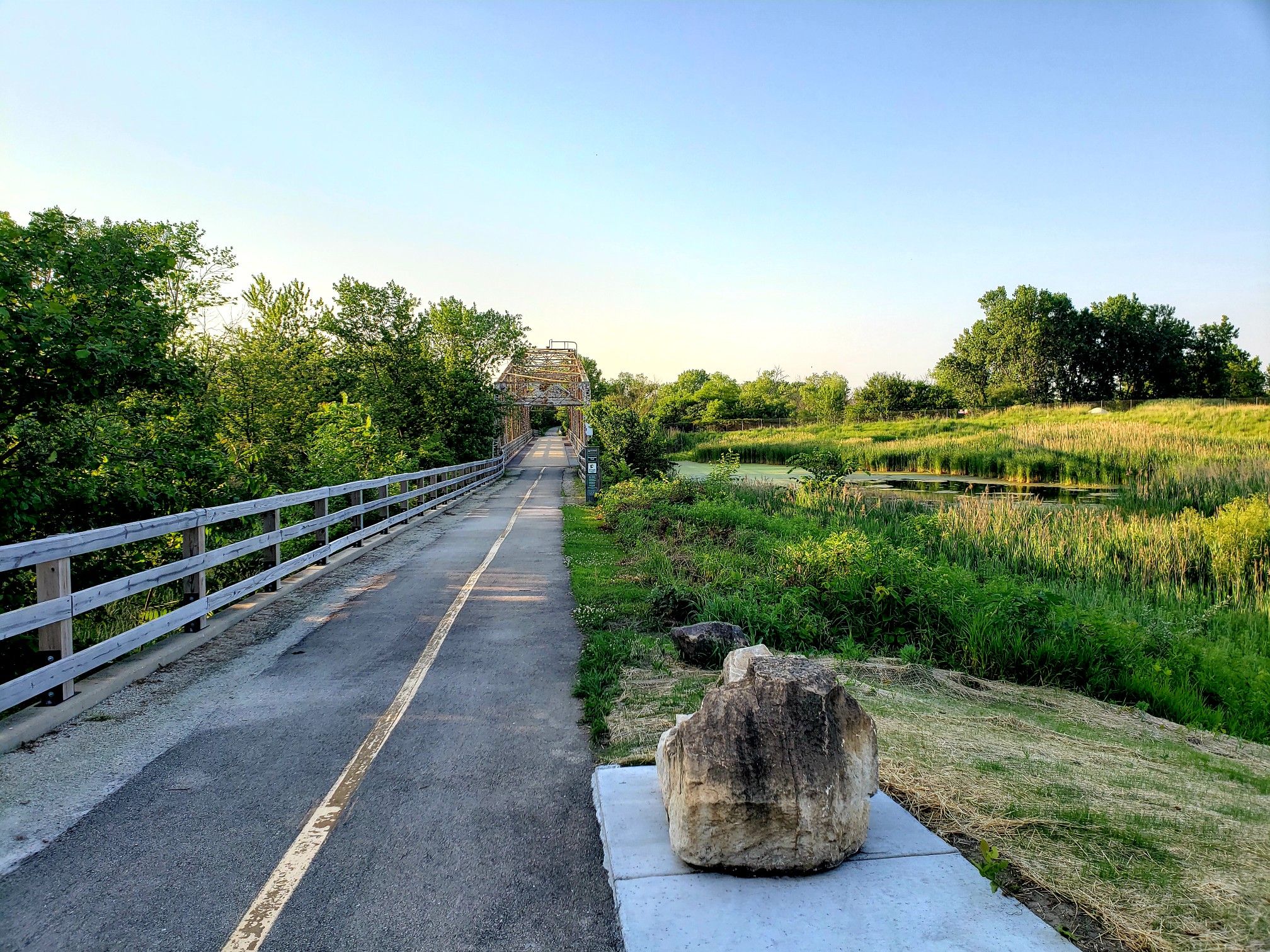 A paved path with a wooden fence leads to a metal bridge at sunset. - Centennial Trail – Prince Pond loop from Fairview Avenue