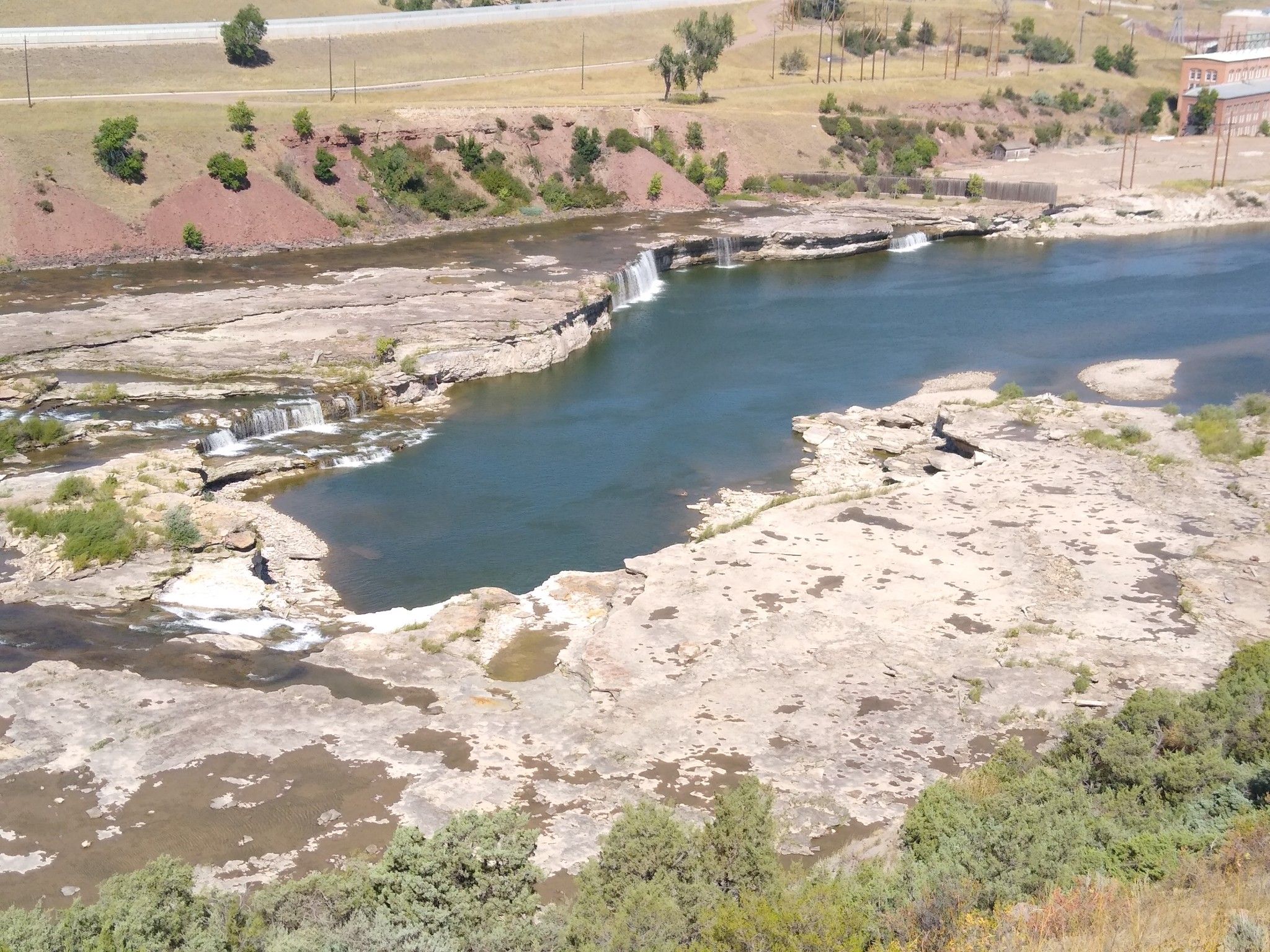 Sunny aerial view of a wide river with rocky banks and small waterfalls. - River's Edge Trail