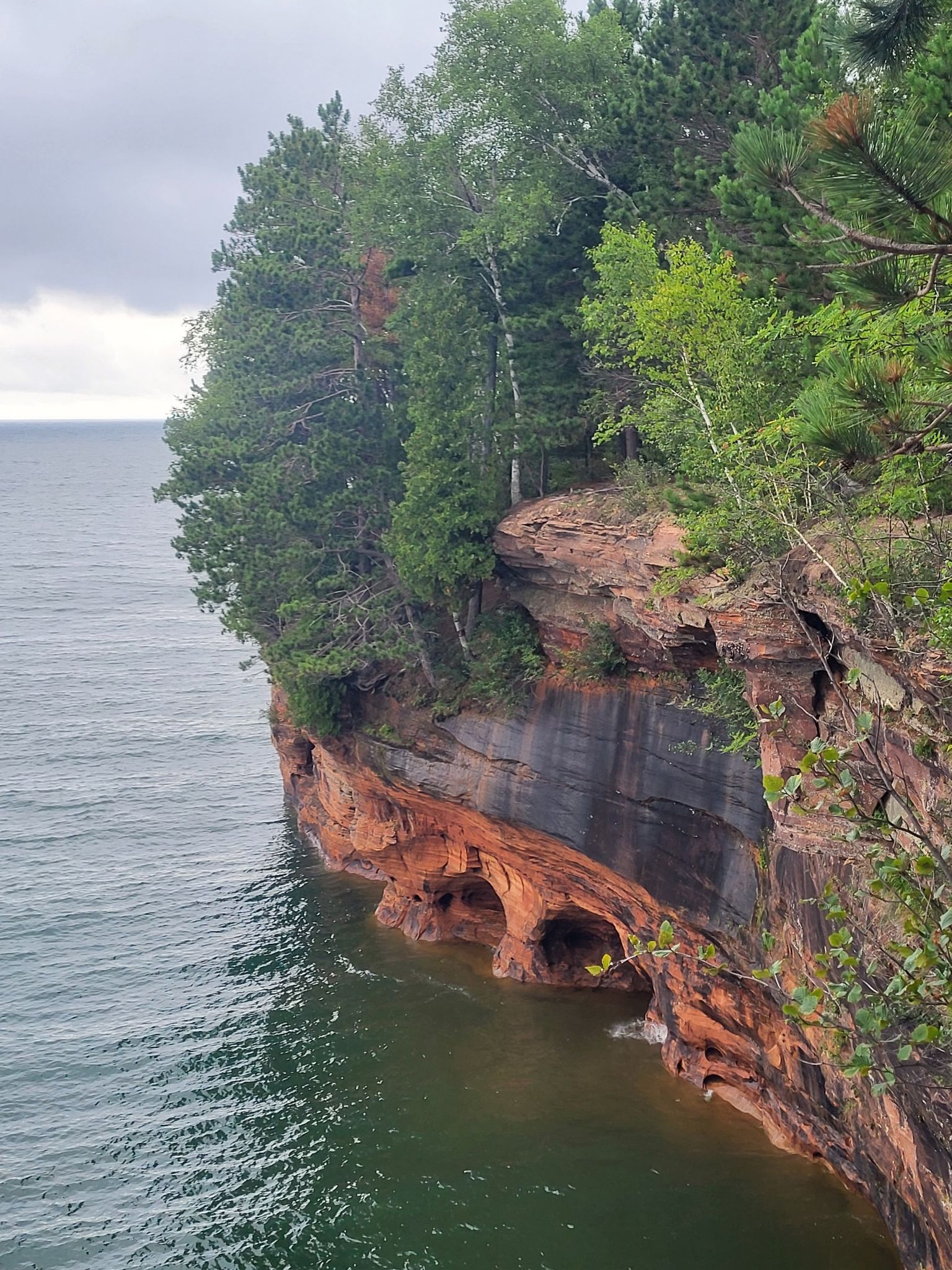 Red rock cliffs with sea caves and green trees over dark water, cloudy sky. - Lakeshore Trail