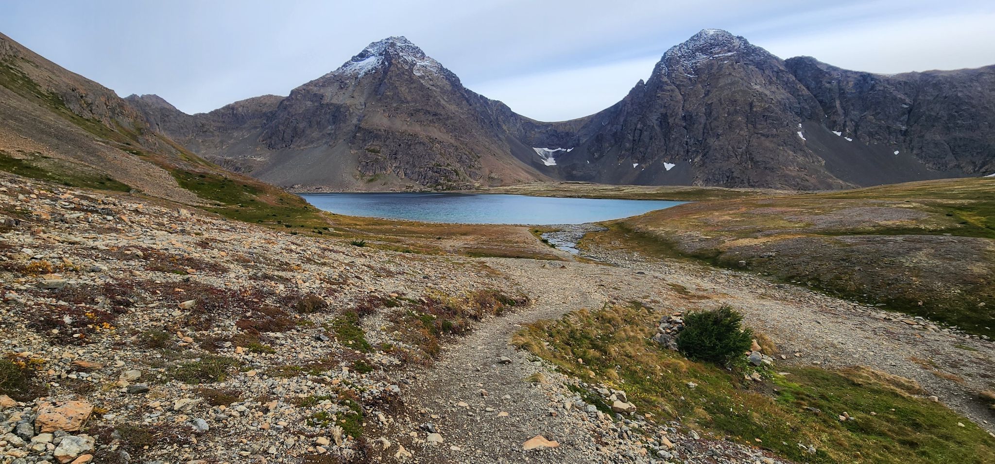 A clear lake between two snow-capped mountains under an overcast sky, with a path. - Rabbit Lake Trail