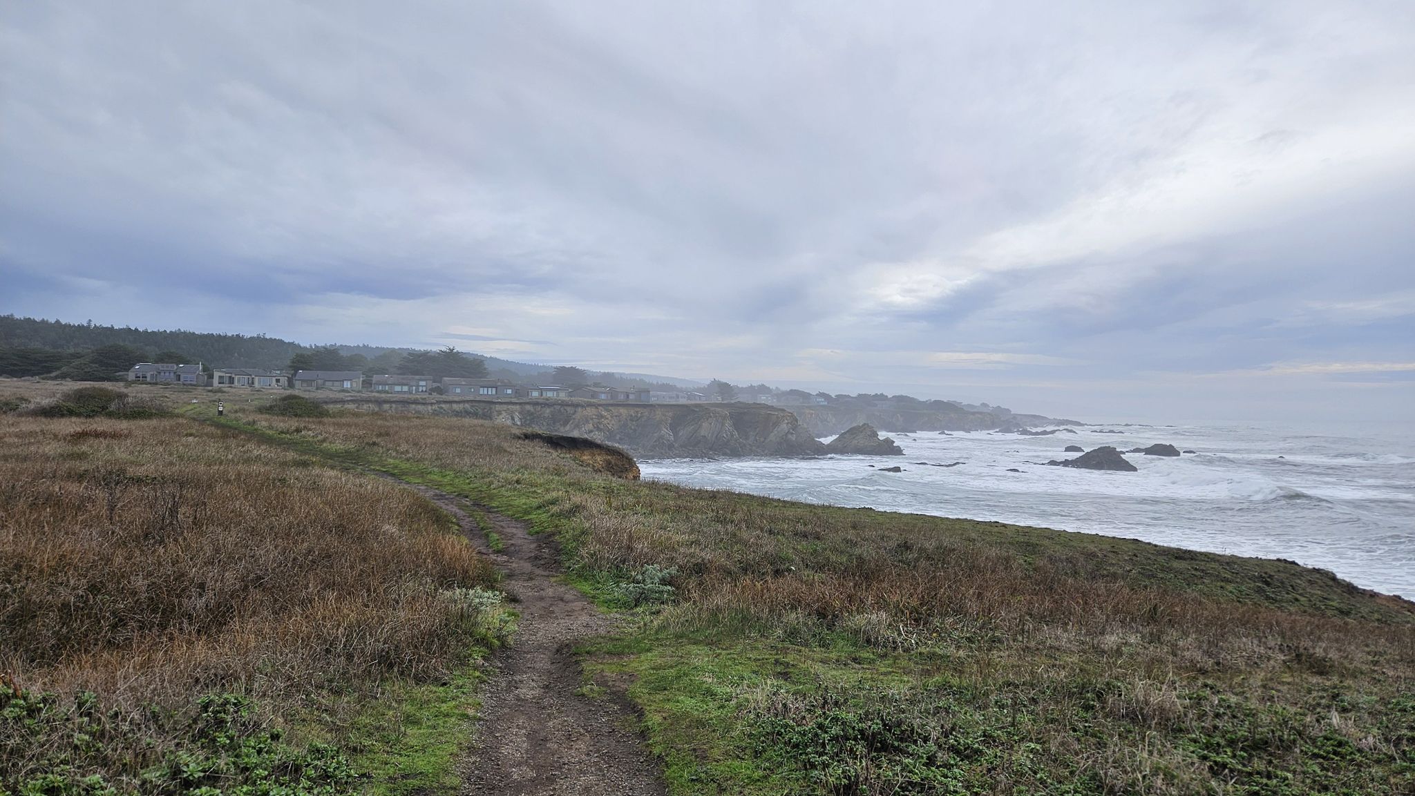 A dirt path on a grassy bluff overlooking a cloudy, wavy ocean. - Bluff Trail Loop