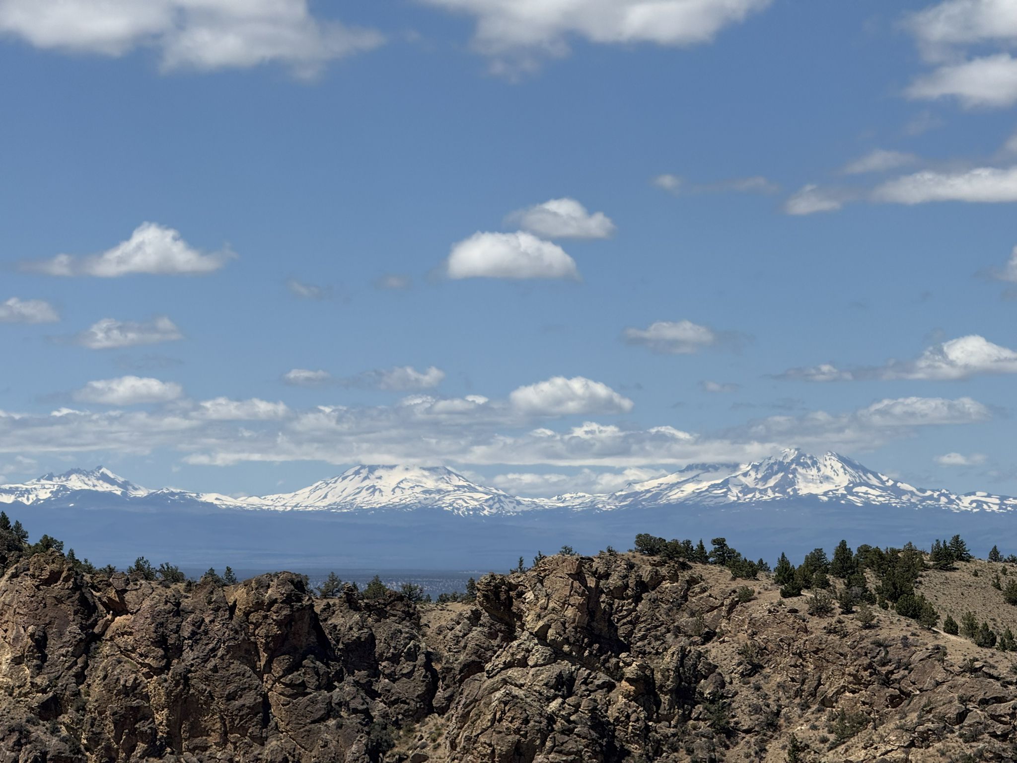 Distant snow-capped mountains rise above rocky hills under a bright, cloudy blue sky. - Gray Butte Trail