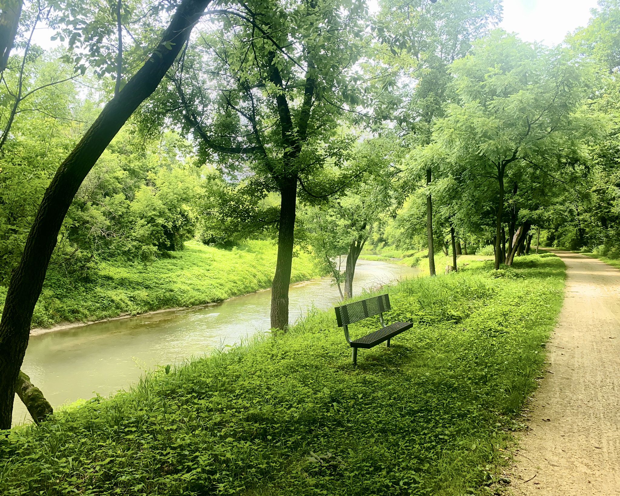 A serene river flows beside a dirt path with a wooden bench. - Heritage Trail Truss Bridge loop from Center Township