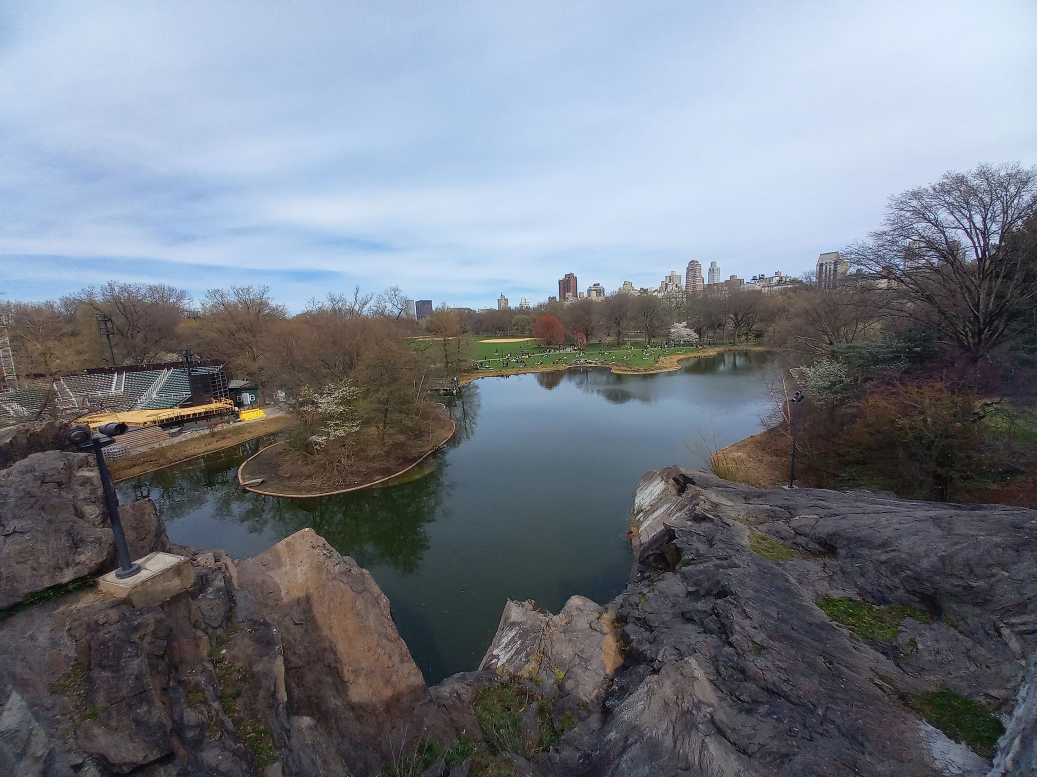 Lake from rocky viewpoint, autumn trees, distant city skyline, cloudy sky. - Central Park Lake and Bow Bridge Loop
