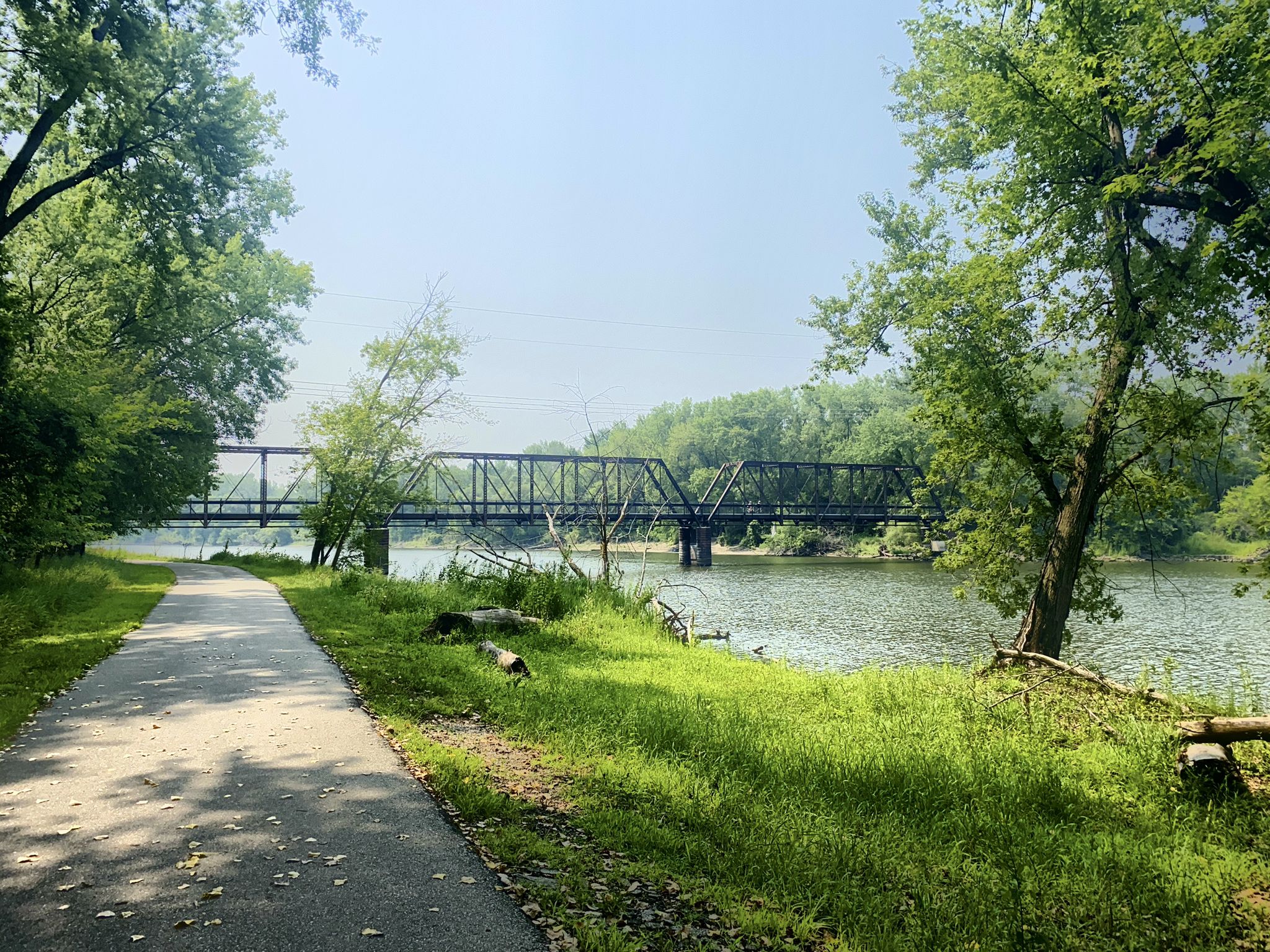 Paved path alongside a river with a long metal bridge, sunny day. - Neal Smith Trail – Des Moines River loop from Staff Parking