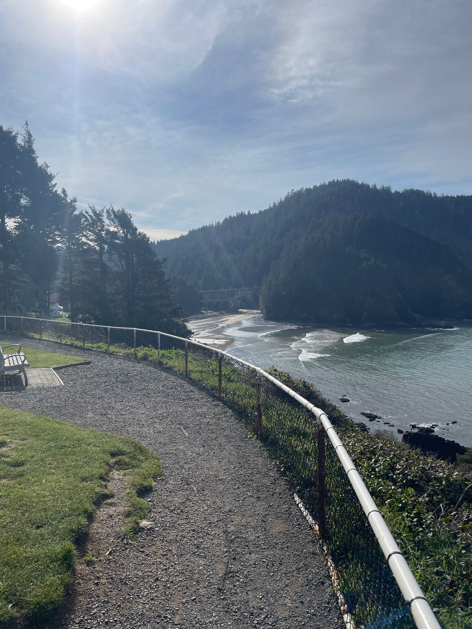 Gravel path with a fence overlooking a bay with waves and forested hills. - Heceta Head Trail