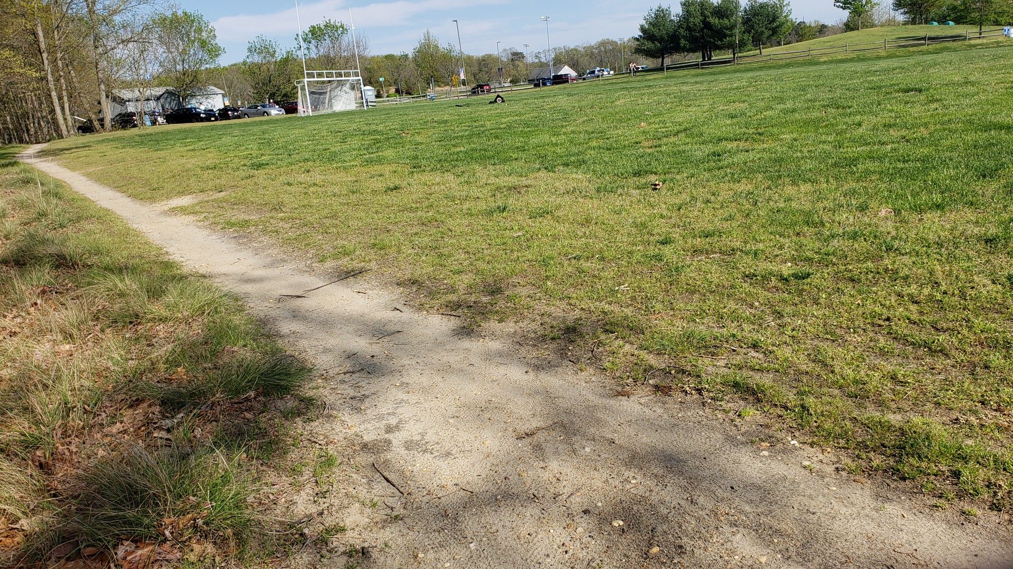 A dirt path curves alongside a bright green grassy field on a sunny day. - Beginner Loop