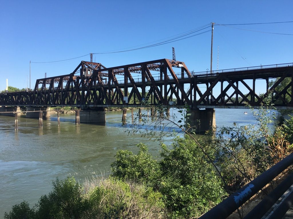 A rusty metal truss bridge spans a wide river with green foliage. - Tower Bridge – Sacramento River Bike Trail loop from John F Kennedy High School