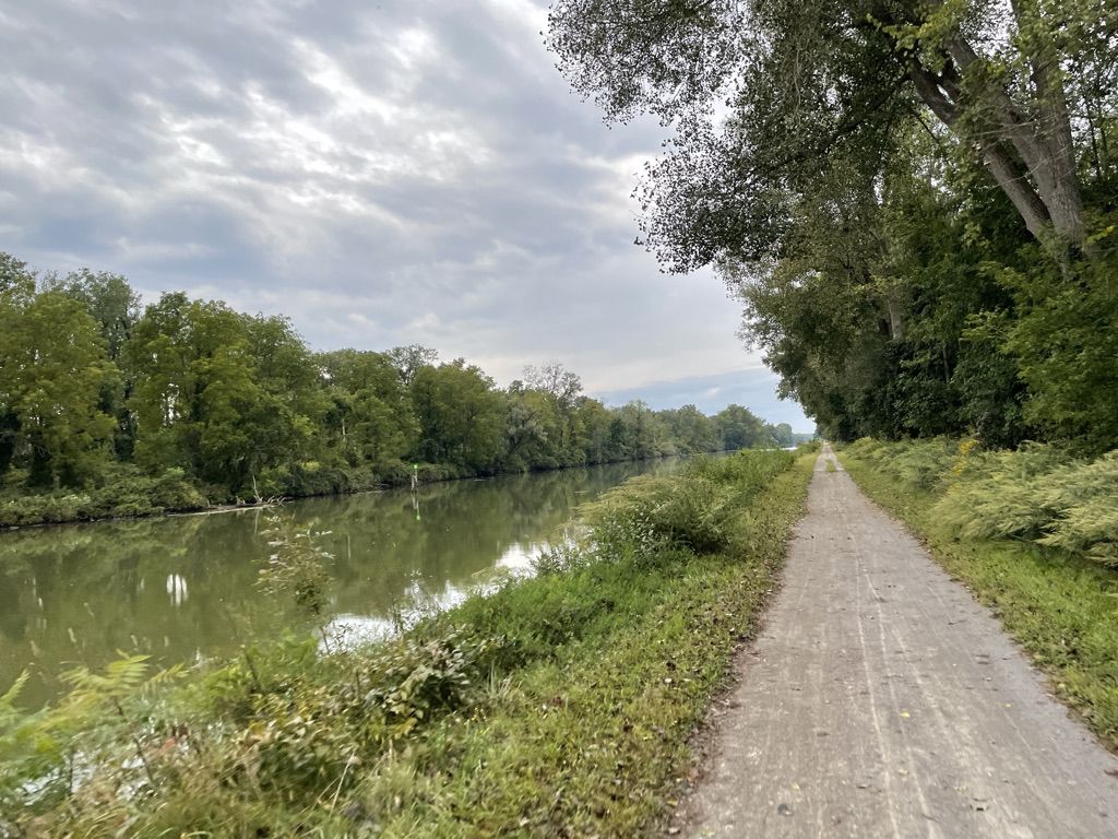 A dirt path runs alongside a river with dense trees under a cloudy sky. - Bridge over the Erie Canal – Fairport NY Erie Canal loop from Wayneport