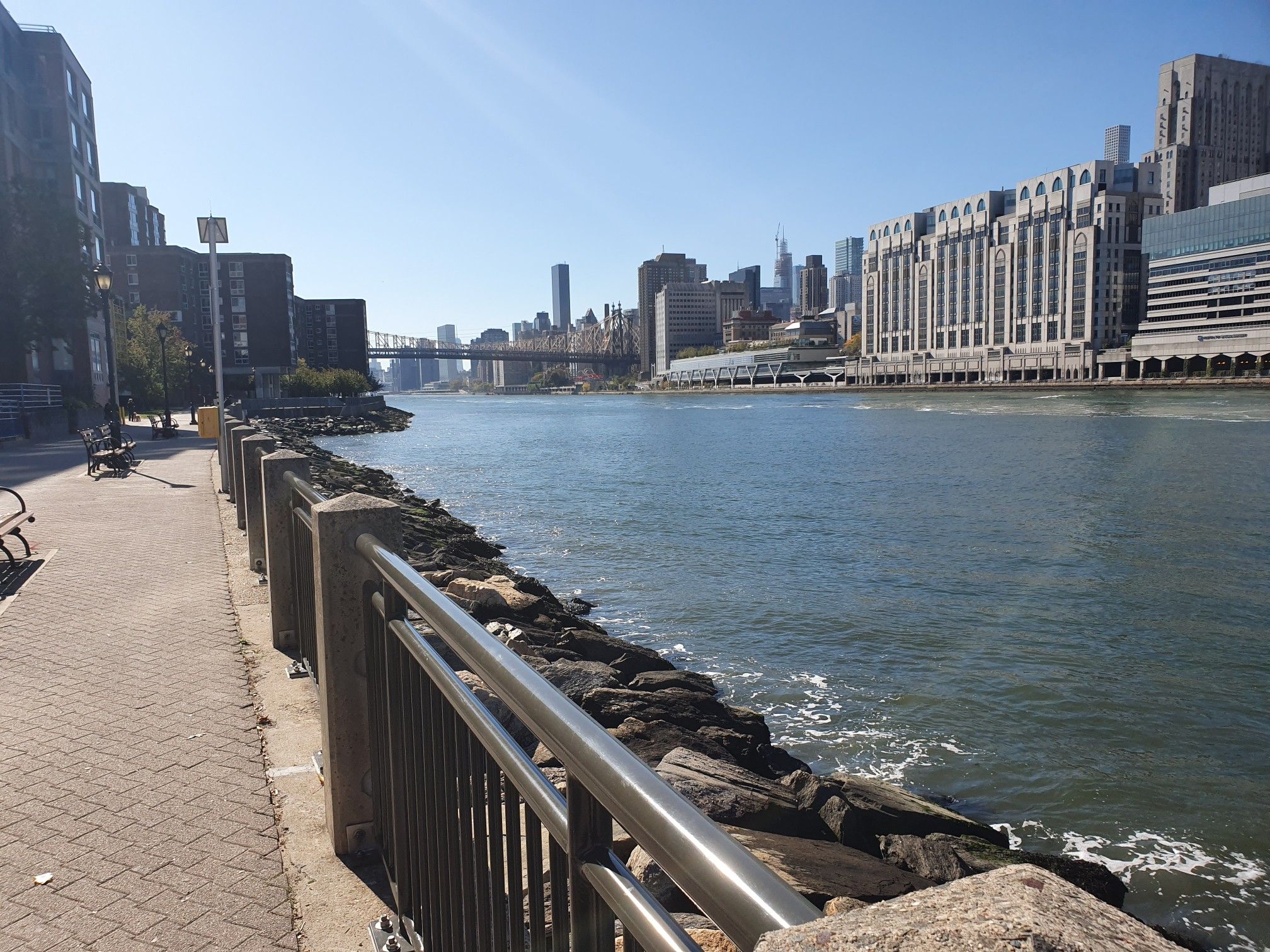 Waterfront path alongside a river with city buildings and a distant bridge. - Roosevelt Island Tramway – Southpoint Park loop from 39th Avenue