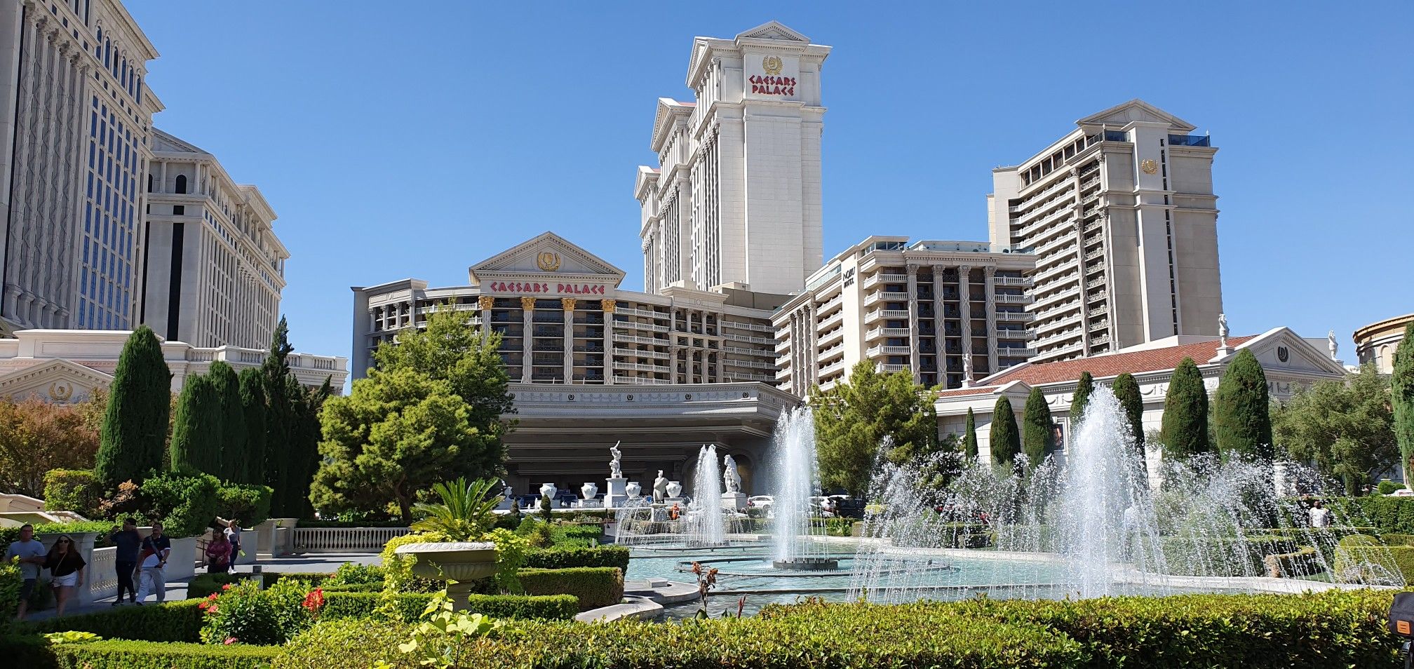 Multiple fountains in a lush garden with grand buildings under blue sky. - The Venetian Resort – Caesars Palace Hotel & Casino loop from MGM Grand