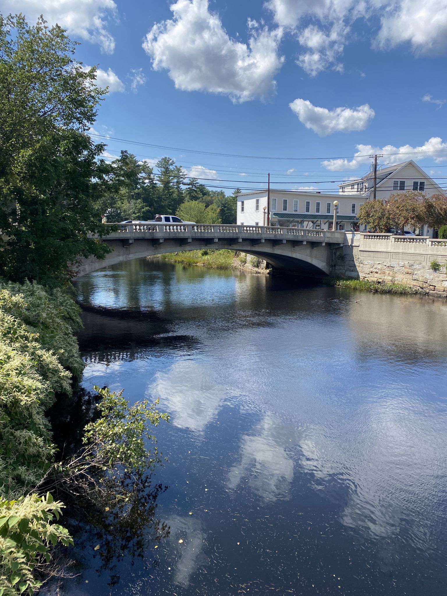 A concrete bridge crosses a river, reflecting clouds on a bright day. - Goffstown Rail Trail