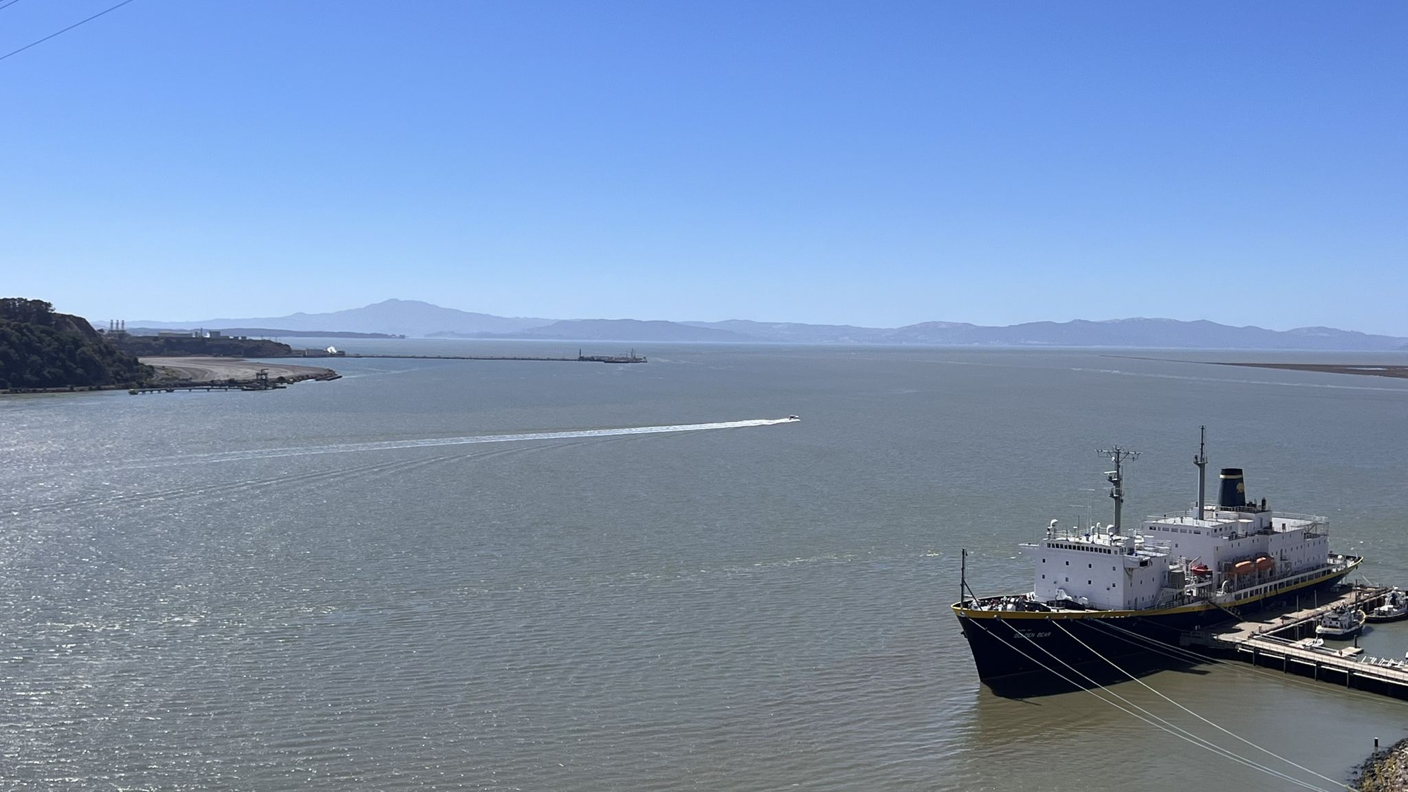 Wide body of water with a large ship docked and a boat leaving a wake. - Alfred Zampa Memorial Bridge – Alfred Zampa Carquinez Bridge loop from California Maritime Academy