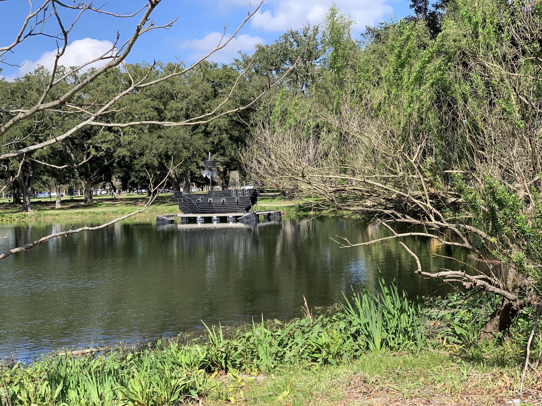 A dark, boat-like structure floats on a pond surrounded by lush trees. - Lafitte Greenway – City Park loop from New Orleans