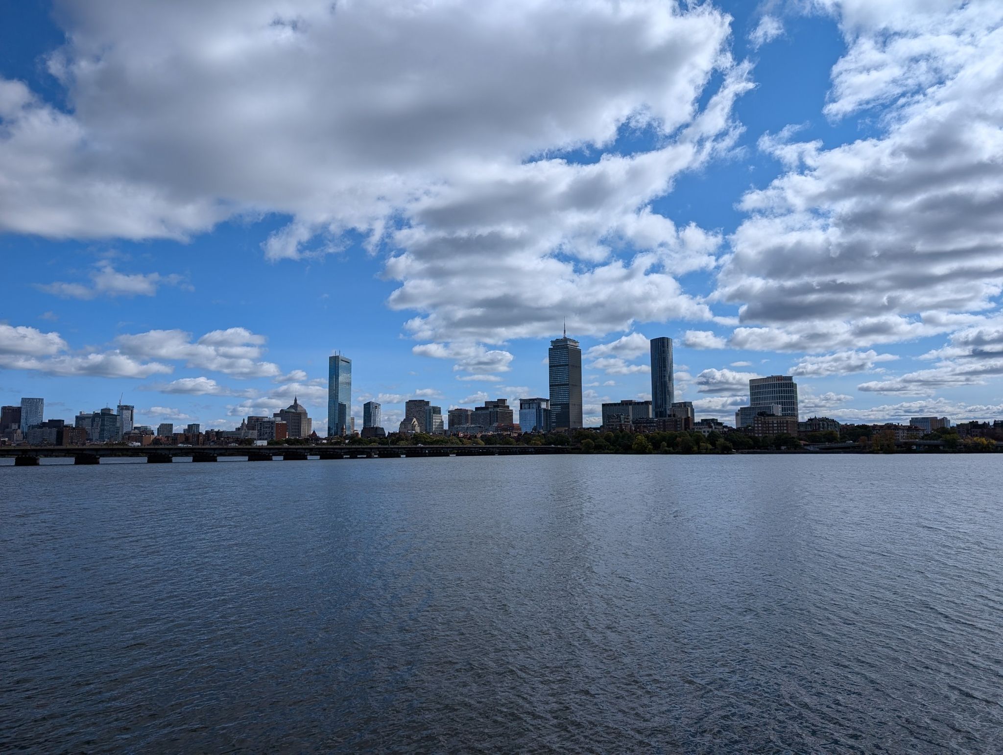 A wide river with a prominent city skyline under a bright blue sky with white clouds. - Harvard Memorial Church – John W. Weeks Footbridge loop from Cambridge
