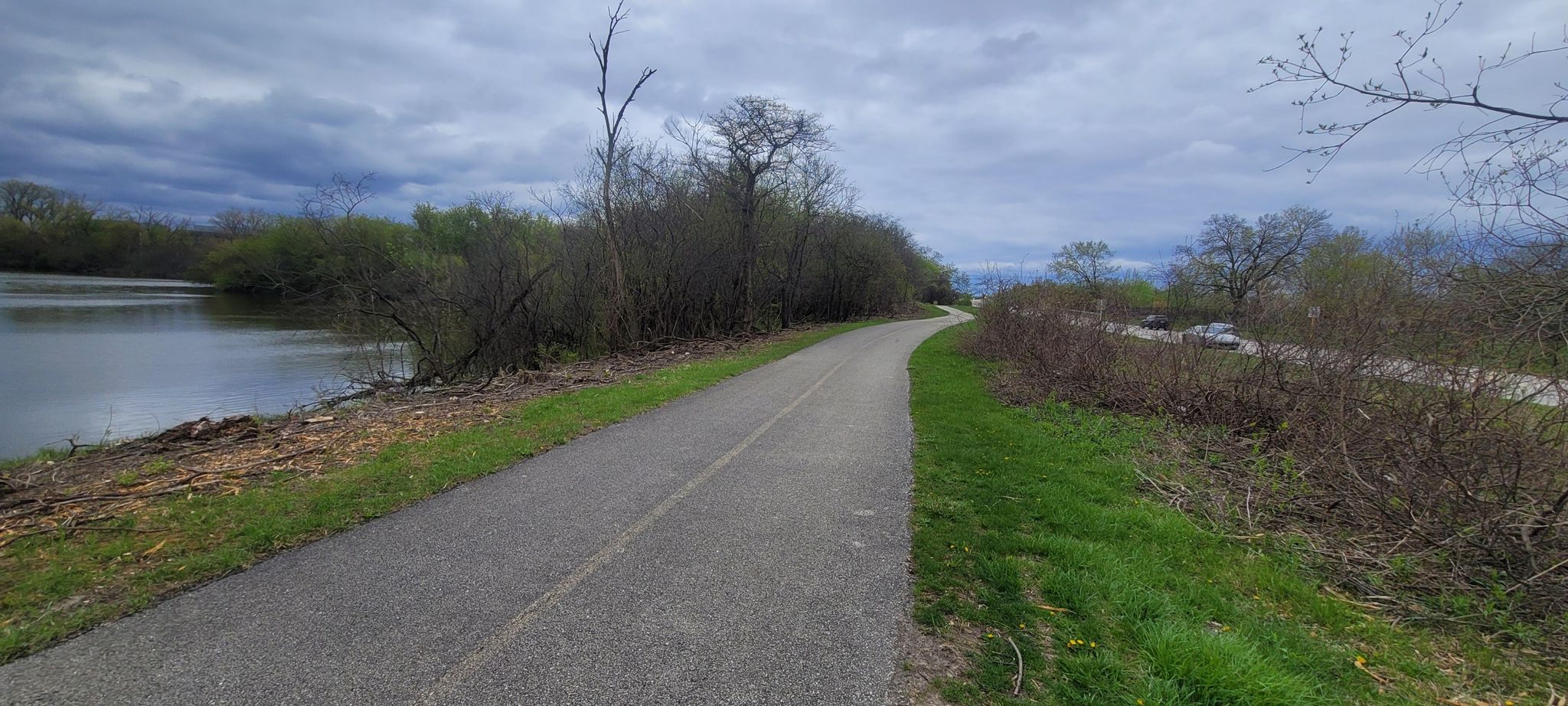 Paved path alongside a calm lake, bordered by trees under a cloudy sky. - North Branch Trail