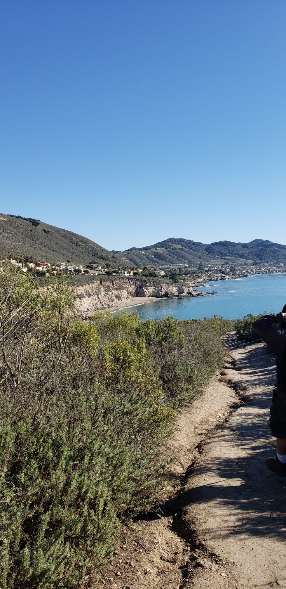 Sunny coastal view with a dirt path, ocean, mountains, and distant houses. - Shell Beach Bluff Trail