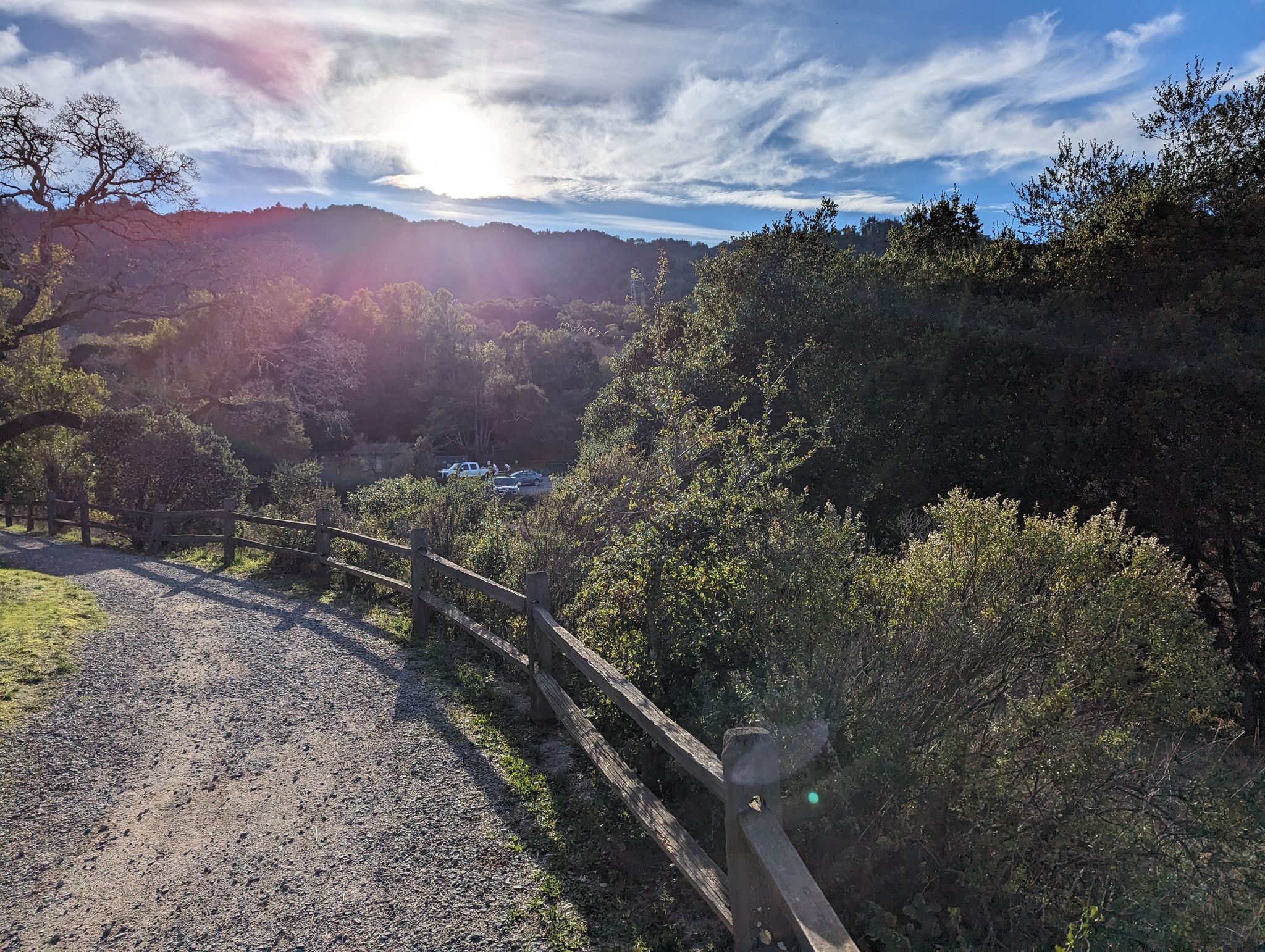 Dirt path with a wooden fence, sun flare over forested hills. - Back Ranch Meadows Loop