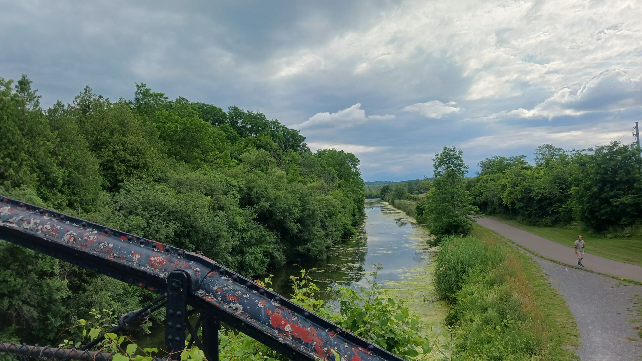 Overhead view of a canal, lush trees, and a person walking on a path under cloudy skies. - Erie Canalway Trail (Syracuse to Dewitt)