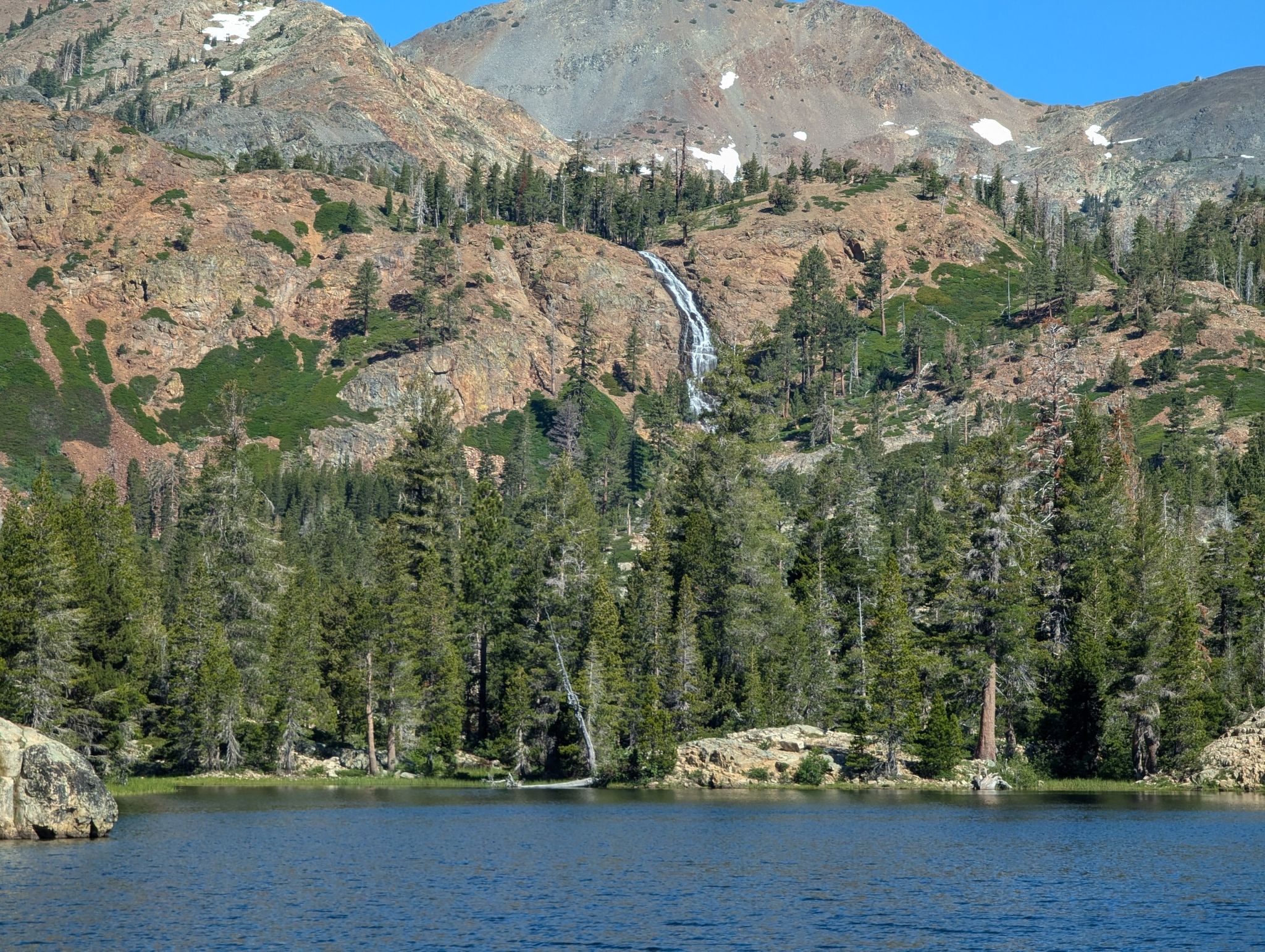 A clear blue lake with pine trees, rocky mountains, and a distant waterfall. - Grass Lake Trail