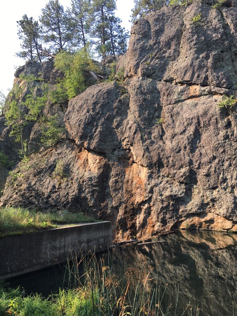 Large sunlit rock formation beside calm water with a concrete structure. - Grace Coolidge Trail