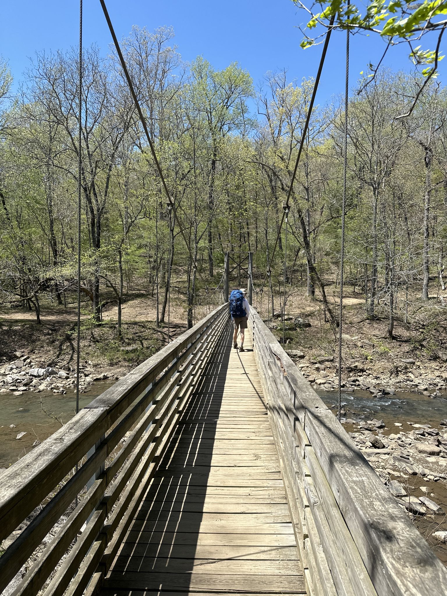 Hiker with a backpack crossing a long wooden suspension bridge over a river. - Suspension Bridge over Lee Creek – Devil's Racetrack loop from Devil's Den State Park
