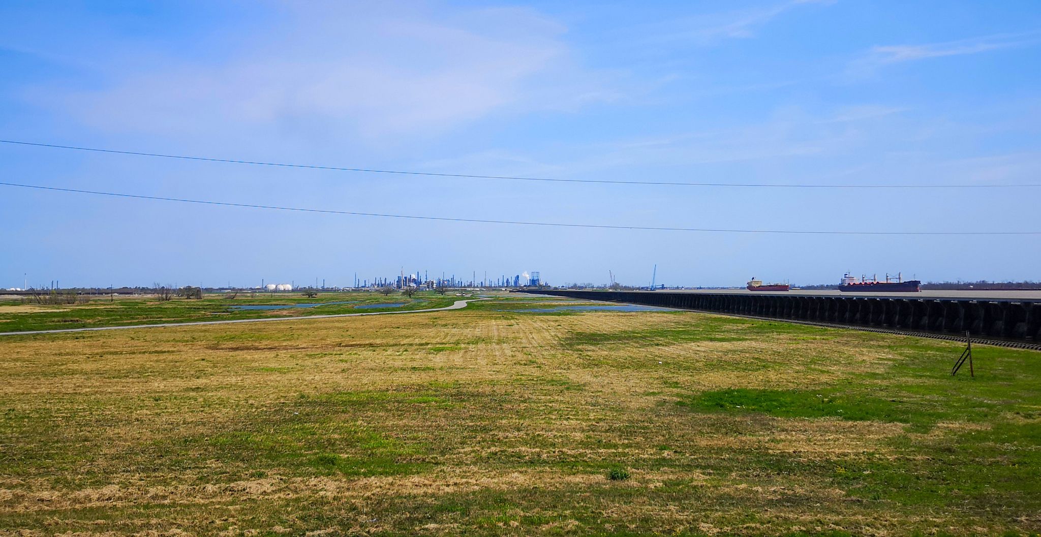 A vast grassy field beside water with distant industrial buildings and ships. - Mississippi River Trail (Louisiana)