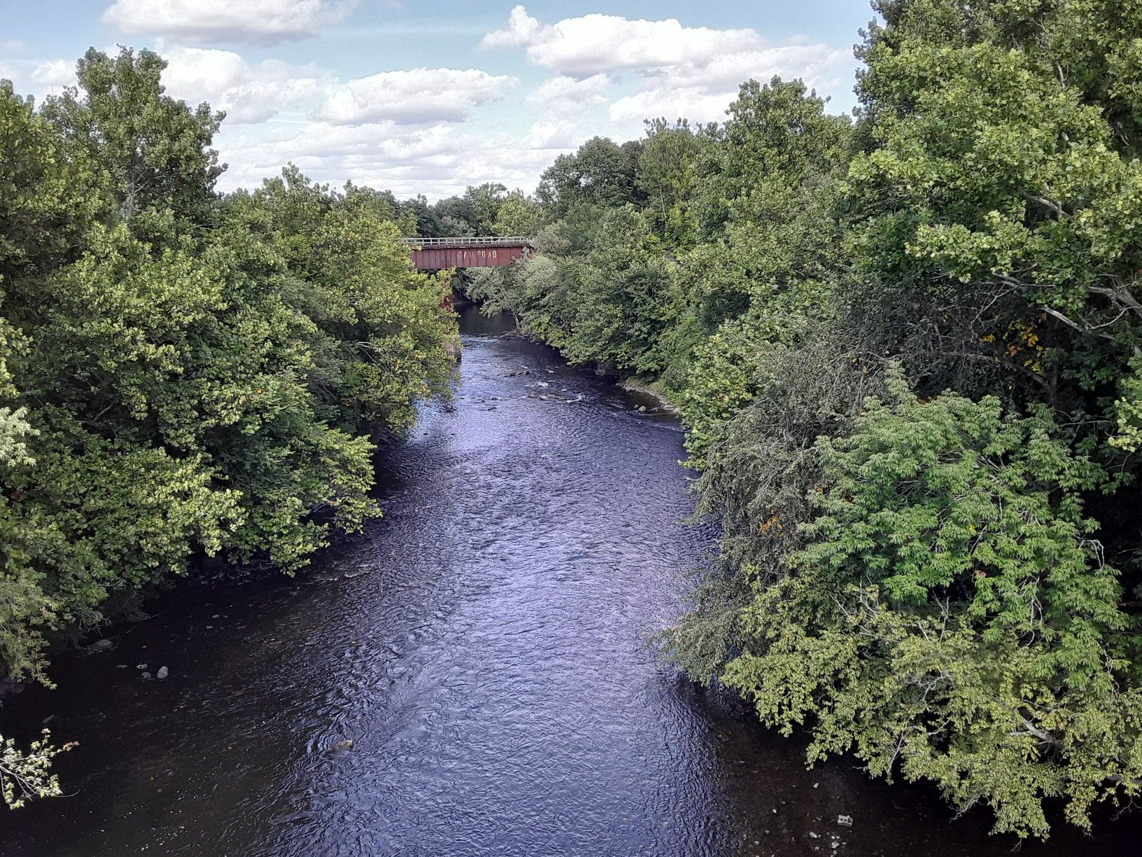 A river flows towards a distant red bridge, surrounded by green trees. - Blackstone River Bikeway – Along the Blackstone River loop from Woonsocket Depot. Polar Express Train Station