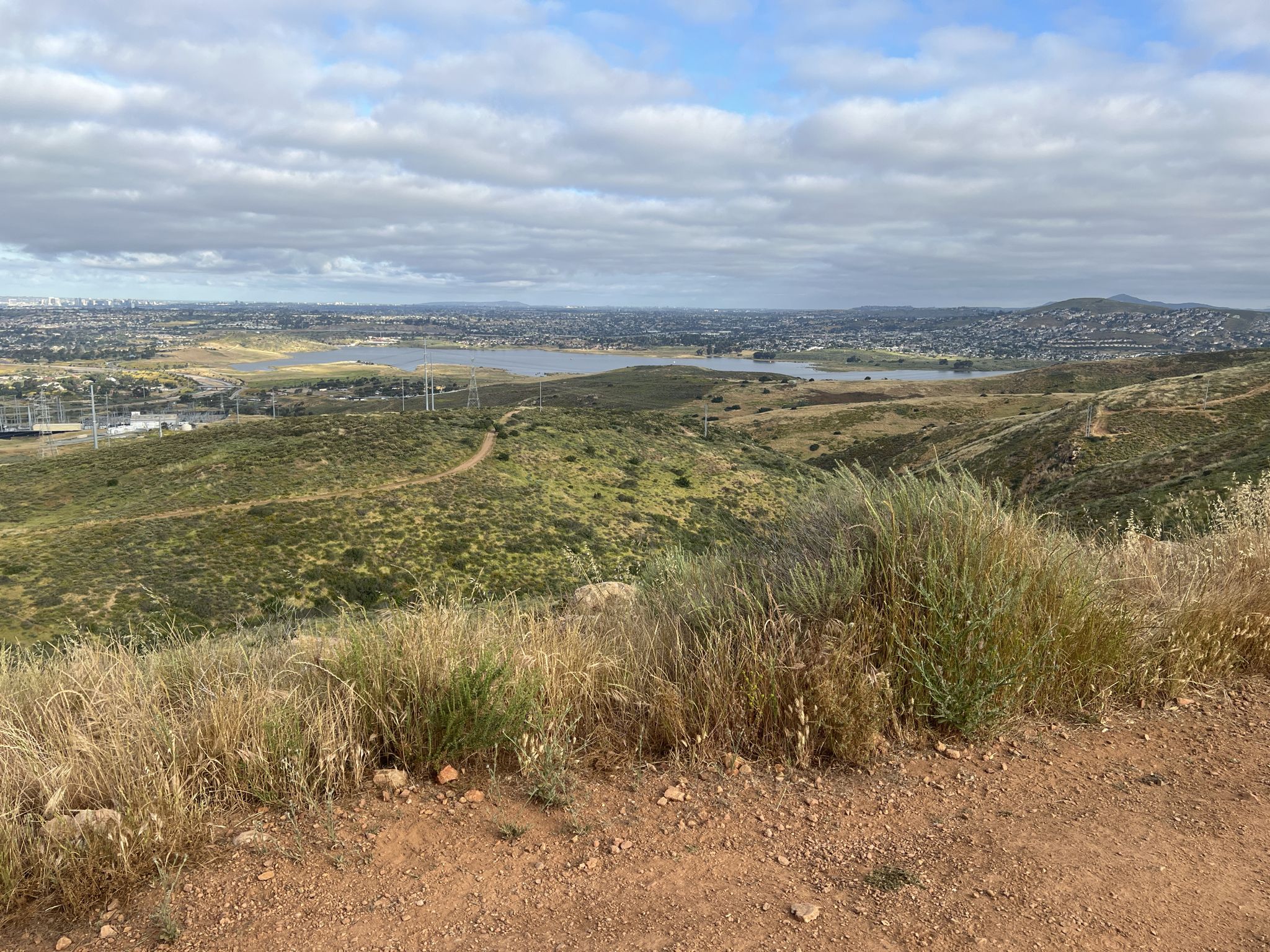 View from a dirt hill over green landscape, distant lake, and city. - Mother Miguel Mountain via Rock House Trail