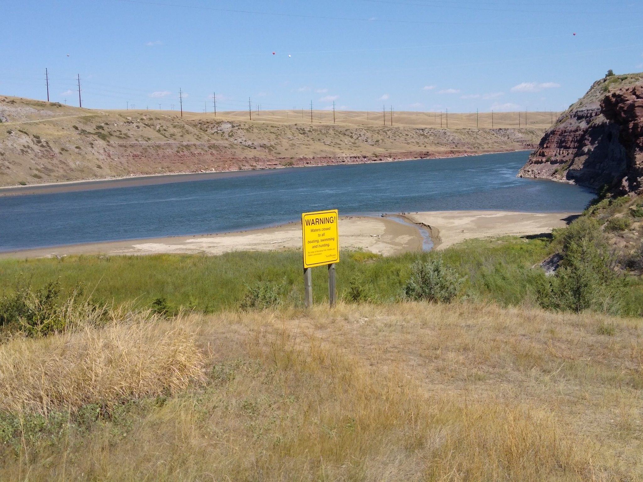 Yellow warning sign on a grassy bank overlooking a wide river. - River's Edge Trail: South Shore