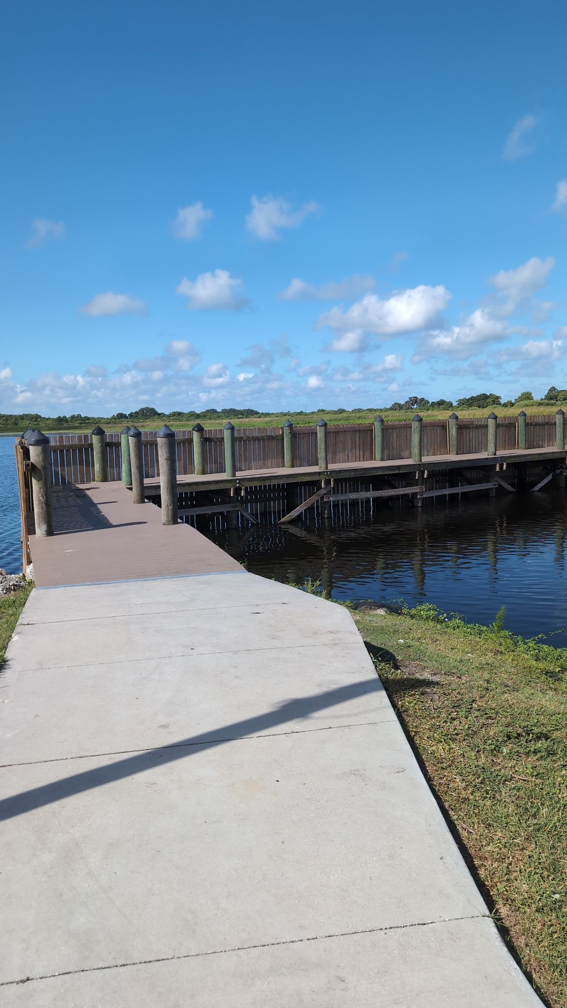 Concrete path leads to a wooden pier over water under a bright blue sky. - Bill Johnston Memorial Pathway