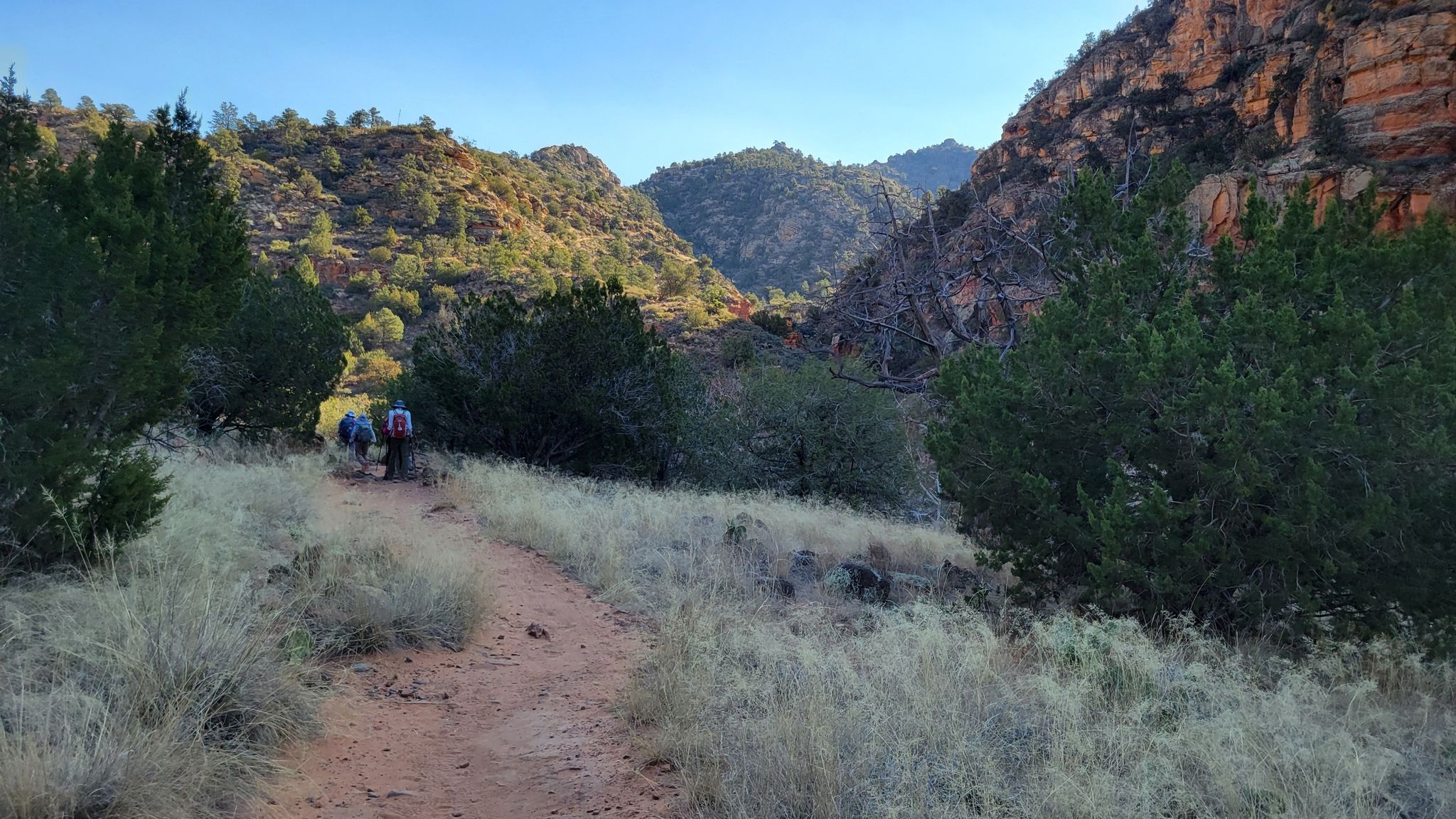Two hikers on a dirt path through a sunny canyon with red rocks. - Bell Trail
