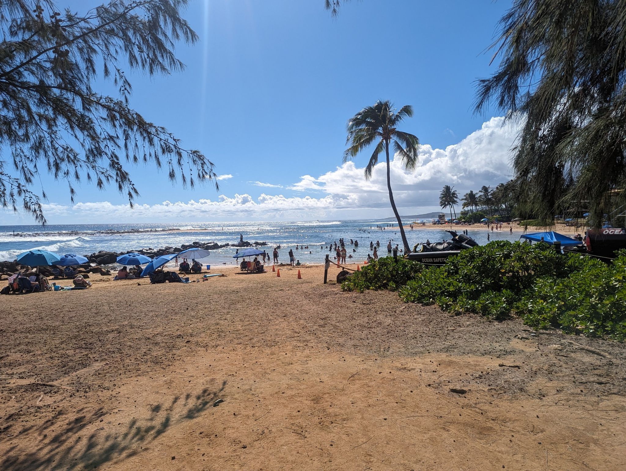 Sunny, crowded sandy beach with people, umbrellas, and palm trees by the ocean. - Spouting Horn Park to Shipwreck Beach