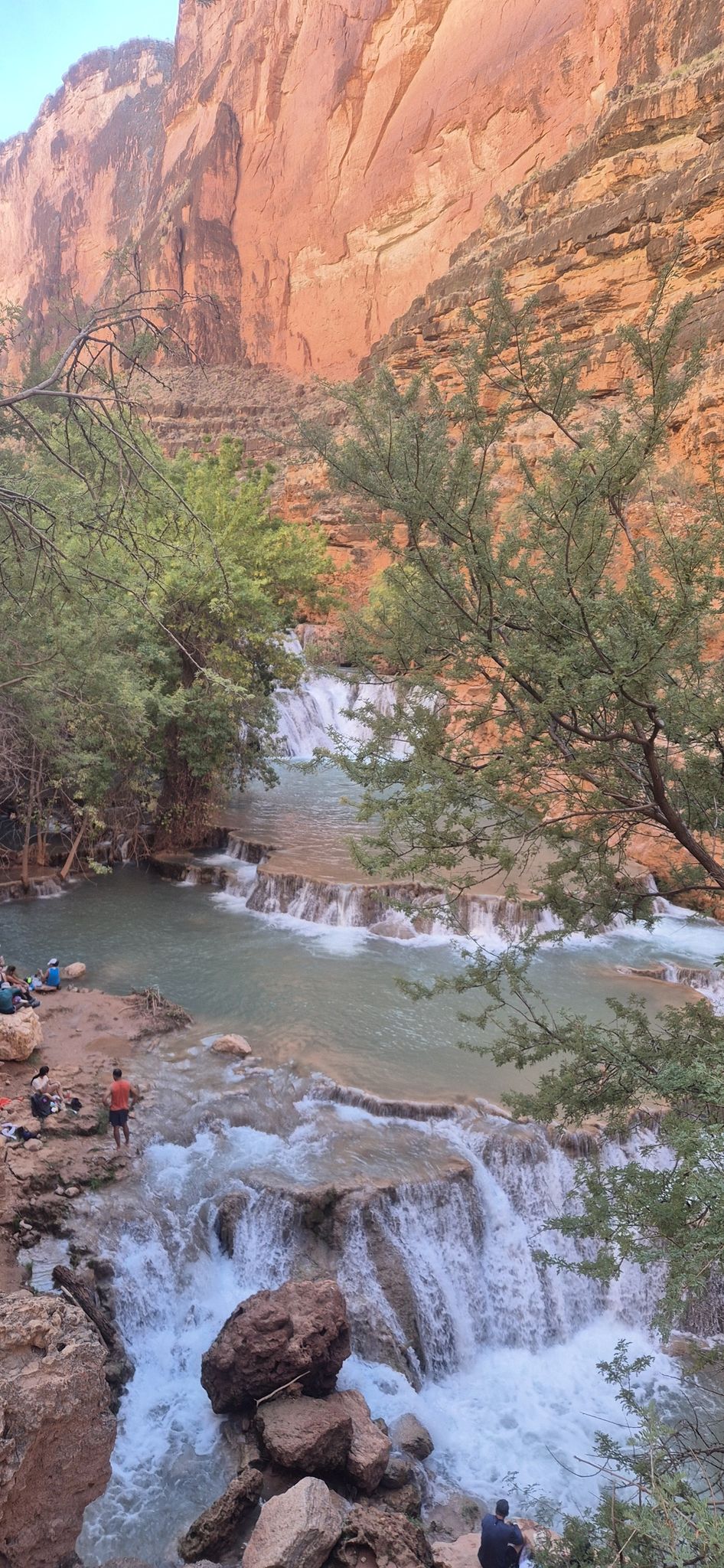People relax by tiered waterfalls in a red rock canyon with green foliage. - Beaver Falls Trail