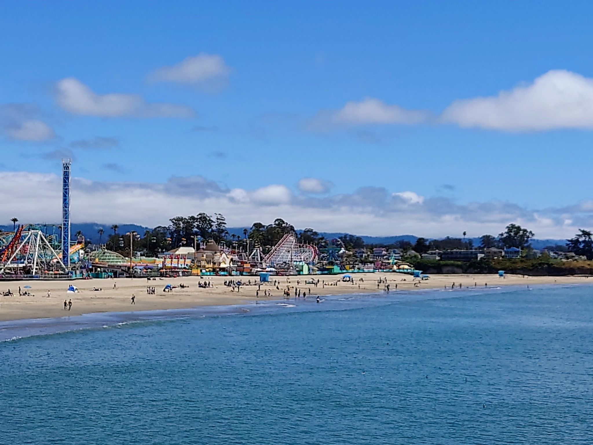 A busy beach with an amusement park and many people under a blue sky. - Santa Cruz Beach Boardwalk – Downtown Santa Cruz Mural loop from Santa Cruz