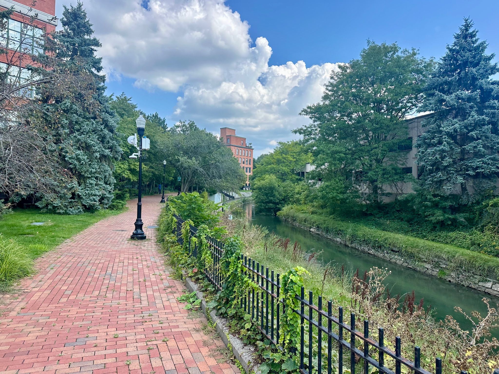 Brick path next to a canal, with green trees and buildings under a partly cloudy sky. - Onondaga Creek – Armory Square loop from Syracuse
