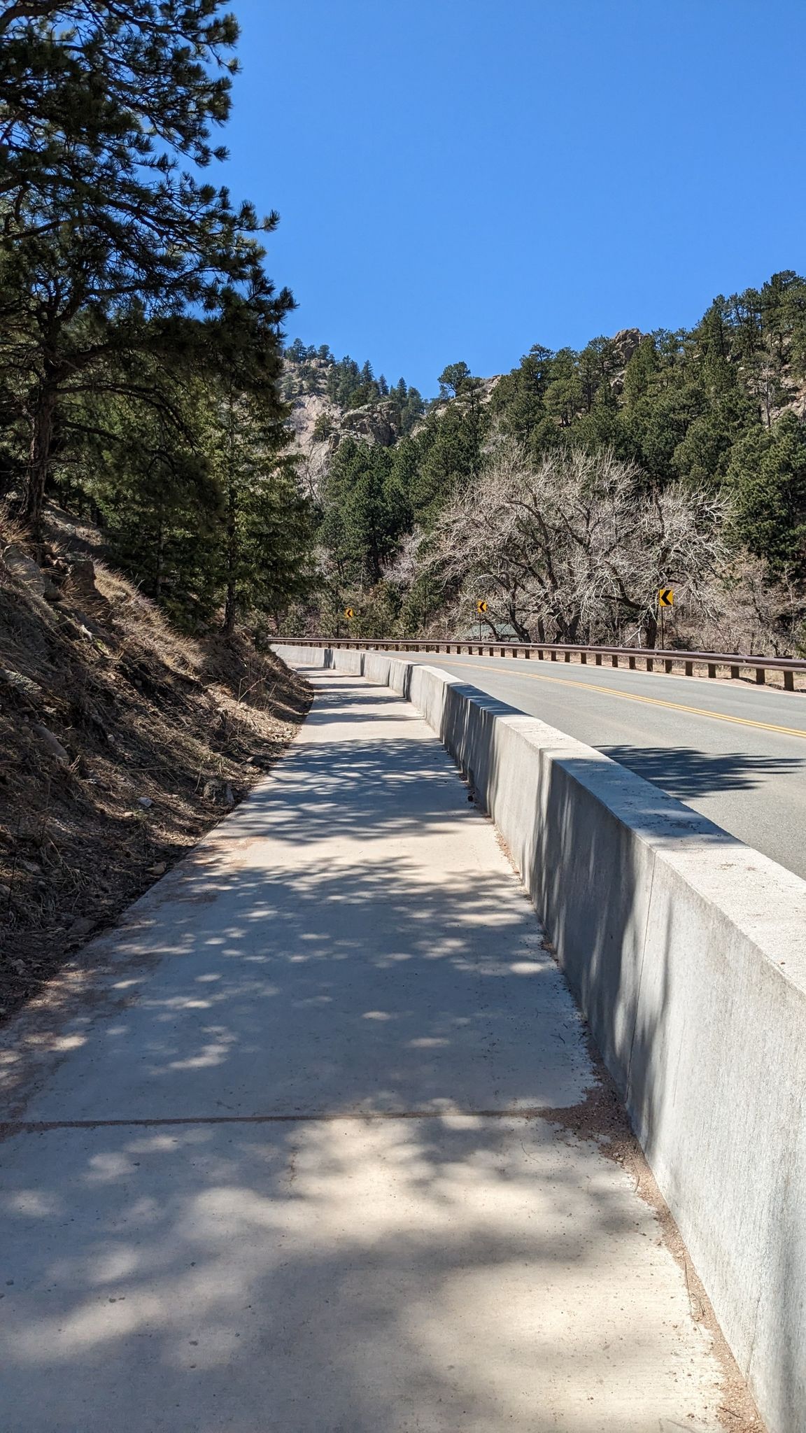 Paved path next to a concrete barrier, alongside a road and forested hillside. - Boulder Canyon Drive
