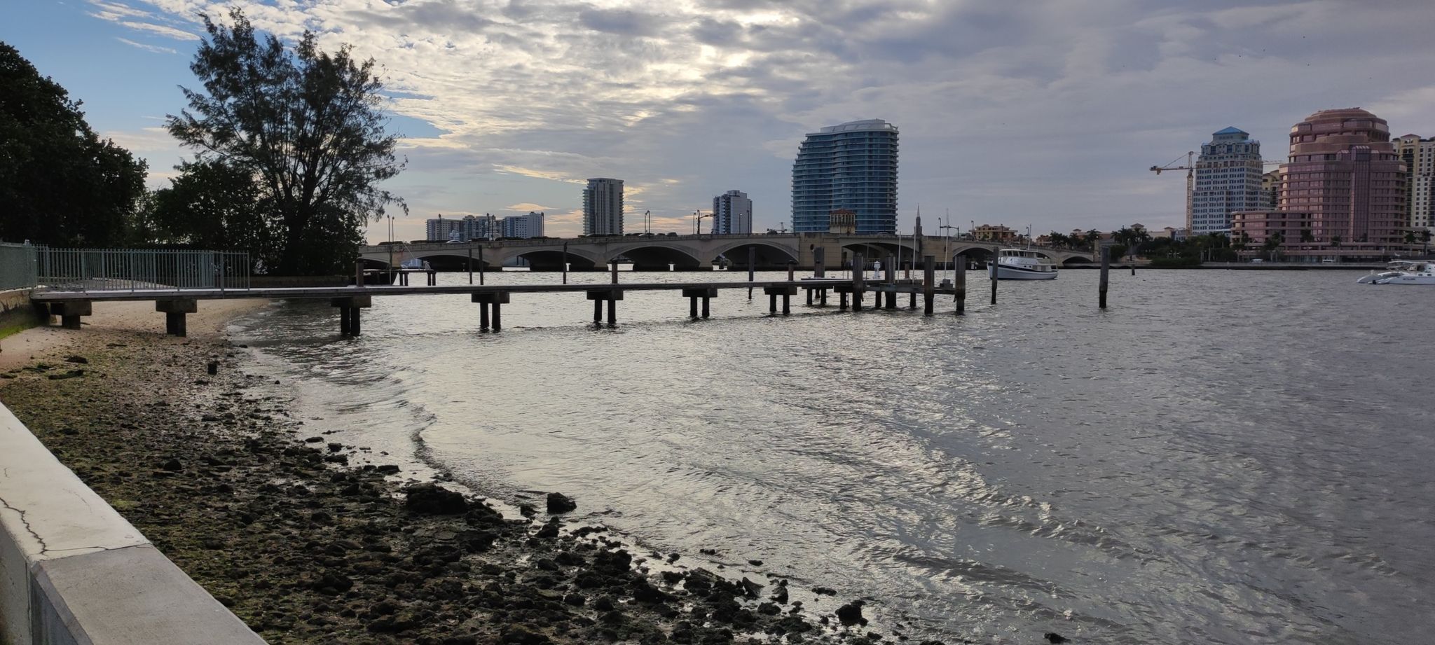 Waterfront with a long pier, city buildings, and a bridge under cloudy sky. - Palm Beach Lake Trail