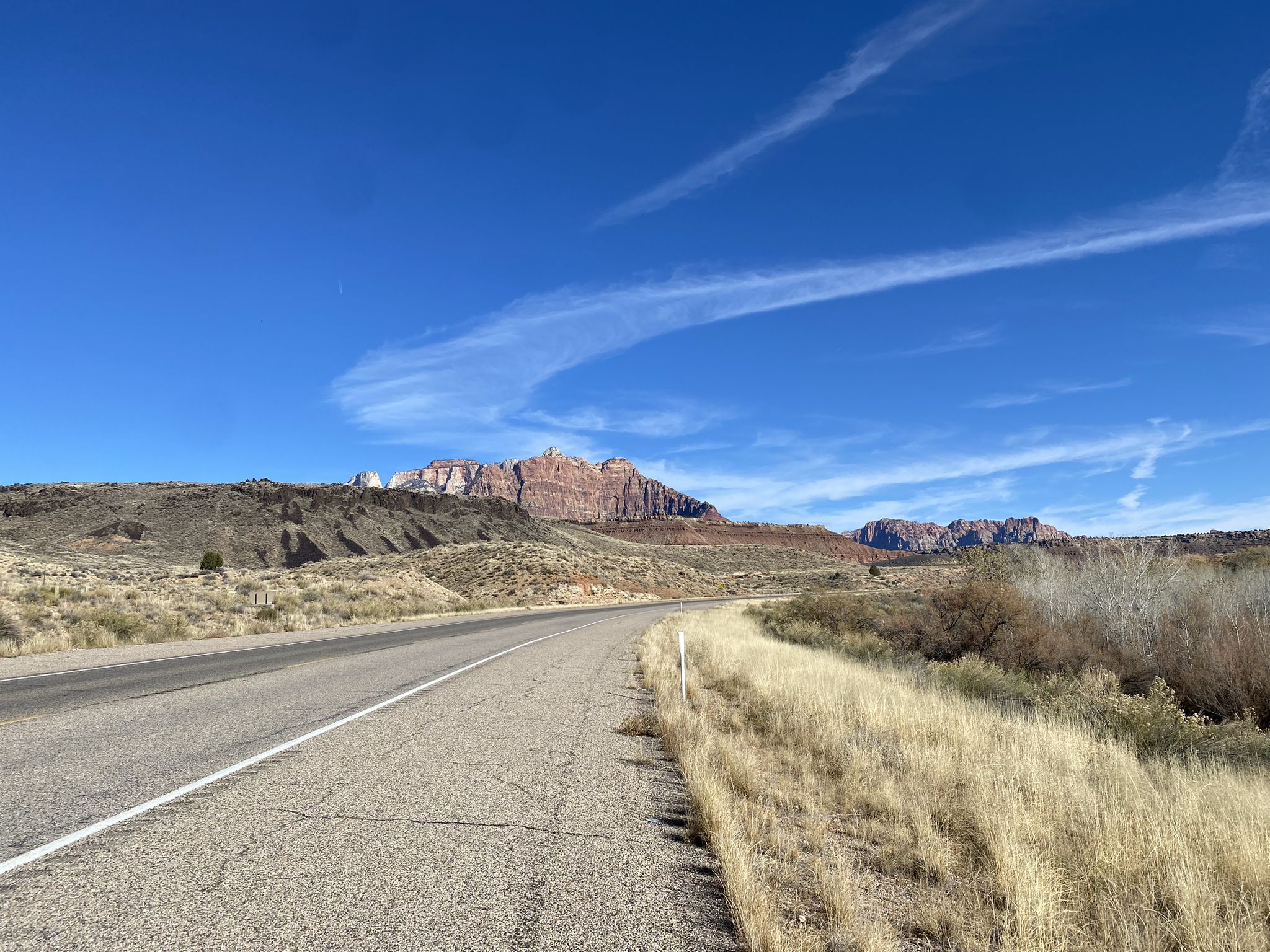 Paved road through arid landscape towards distant red rock mountains under clear sky. - View to South Mountain – Majestic View Lodge loop from Springdale
